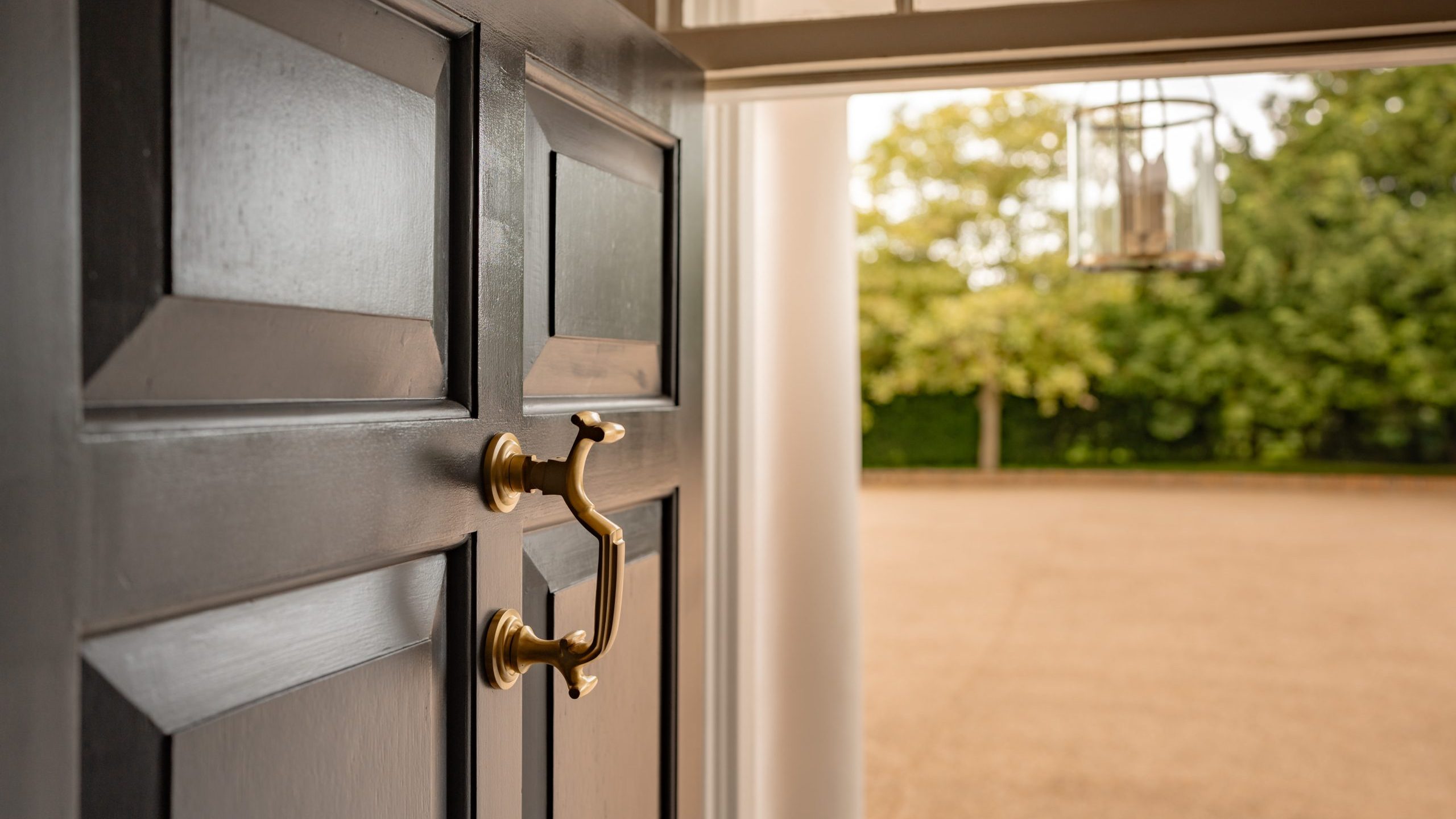 A brass doorknocker and handle are affixed to a partially open dark wooden door, revealing an outdoor scene with bare pavement and grassy trees in the background. A hanging lantern is visible in the open doorway.