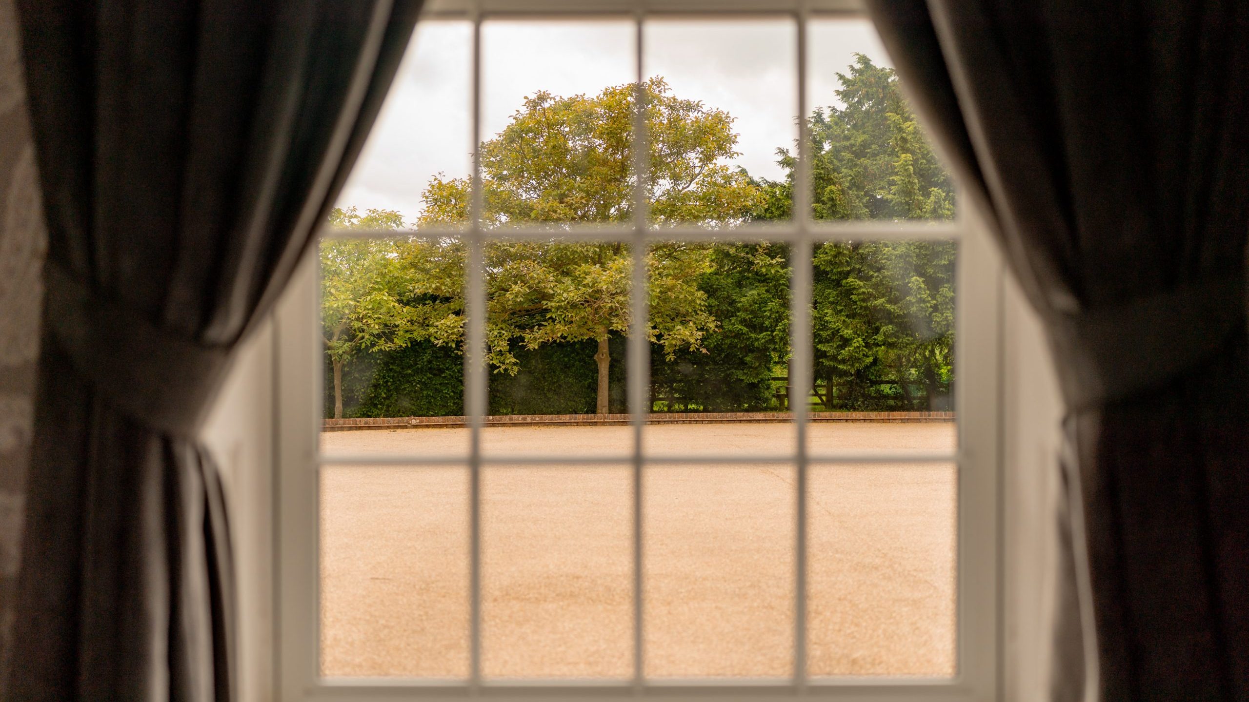 A sunny outdoor scene viewed through a window framed by dark curtains. The window design has white grid lines dividing the glass into small panes. Outside, there is a courtyard bordered by trees, with green foliage highlighted by the sunlight.