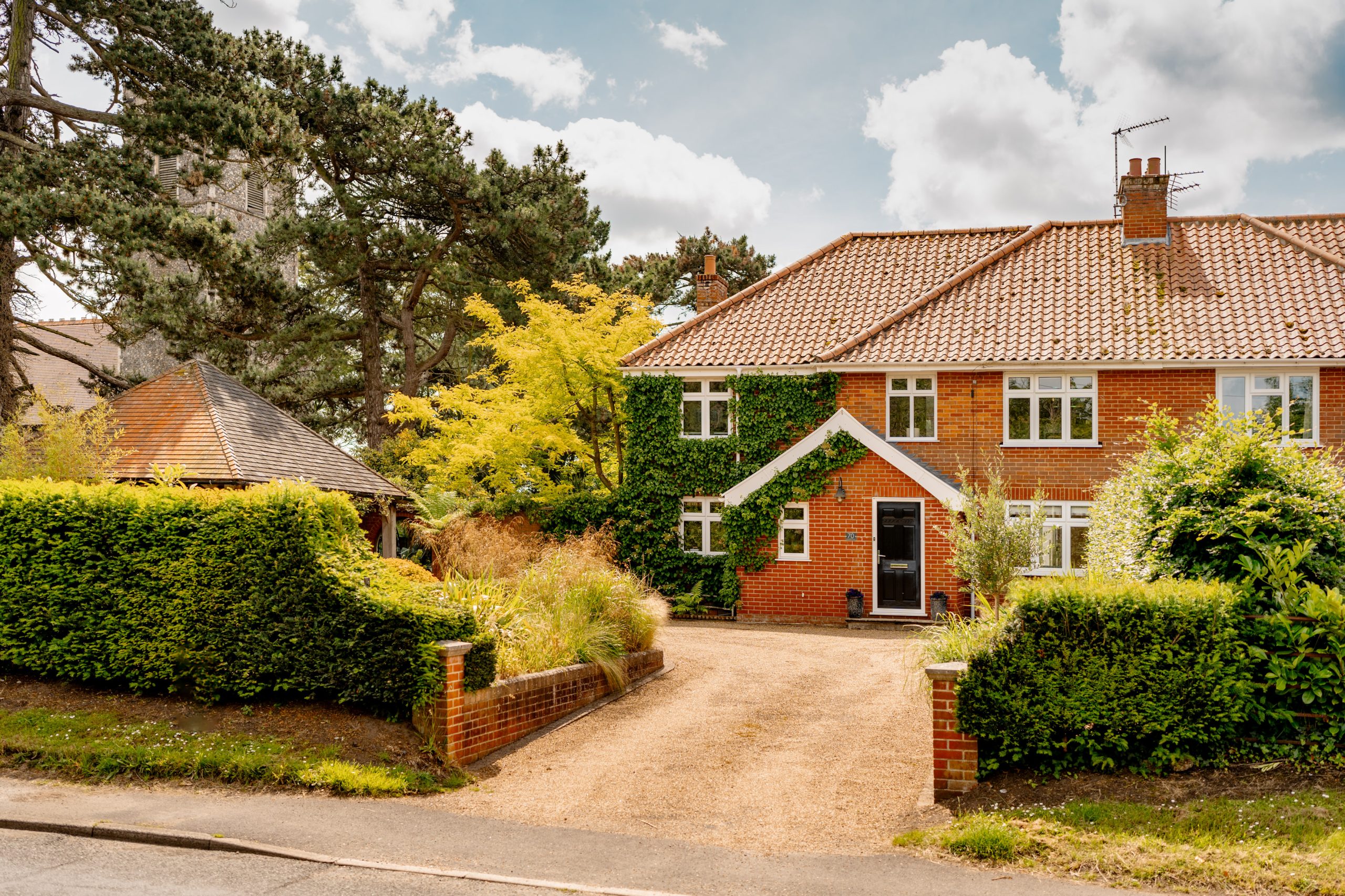 A charming two-story red brick house with ivy growing on part of the facade, featuring a red-tiled roof and dormer windows. It has a gravel driveway, well-maintained hedges, and trees in the yard. The sky is partly cloudy with a mix of sun and clouds.