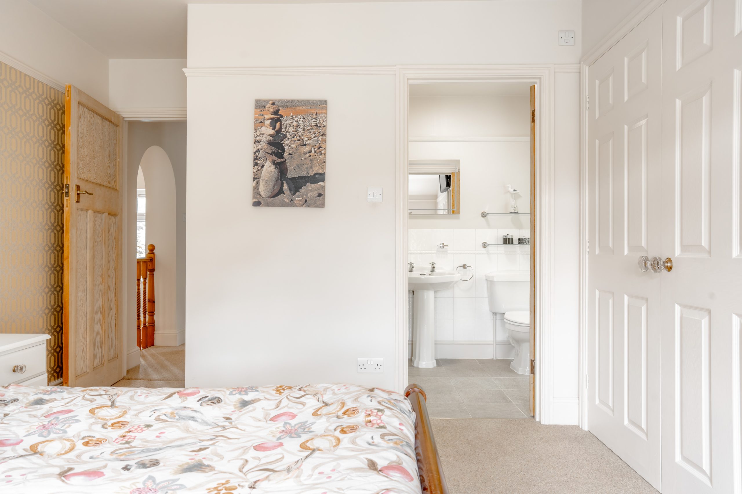 A bright, minimal bedroom with white walls featuring a double bed with a floral duvet cover. The room has a wooden door leading to a hallway, and a door to a bathroom with white fixtures. A photo of stacked stones hangs on the wall.