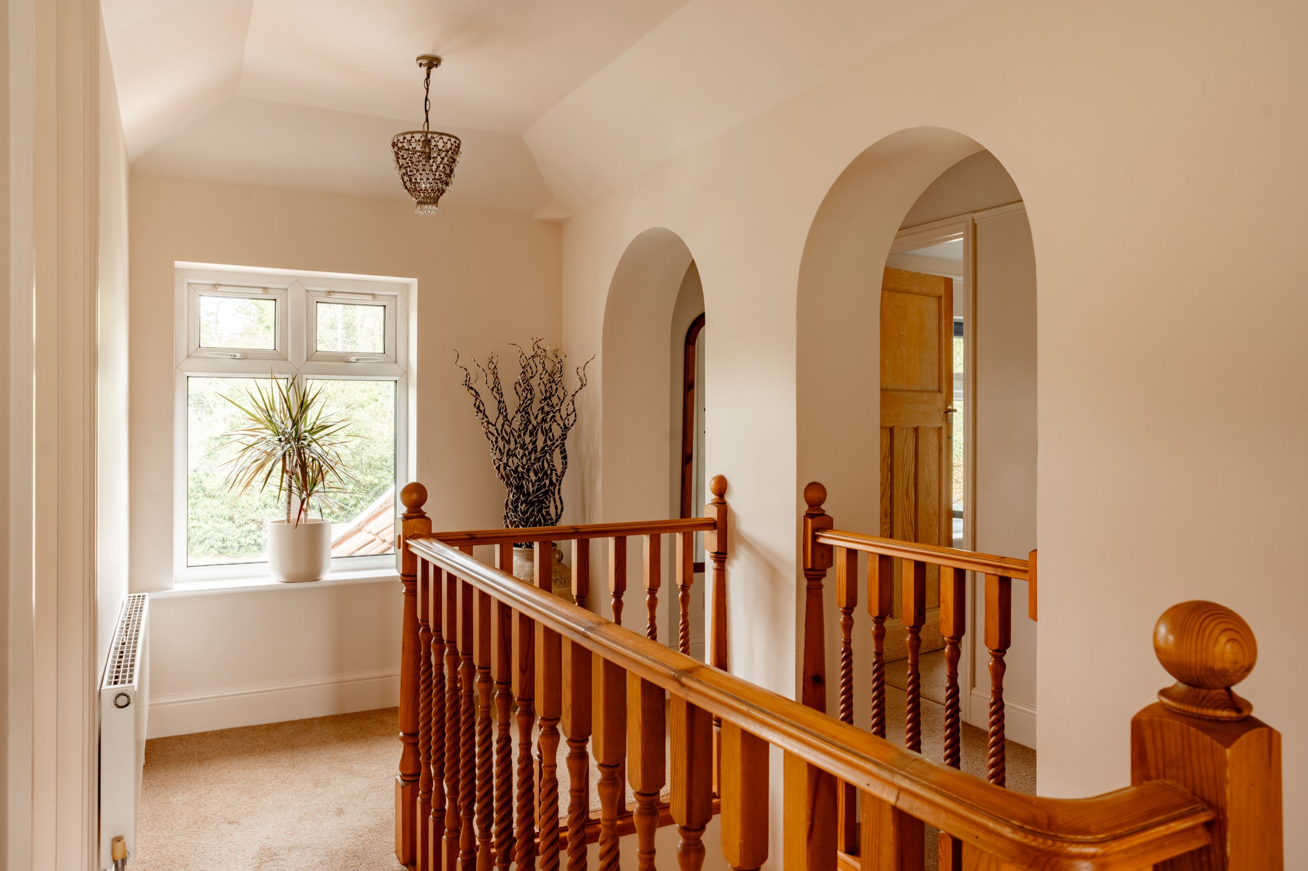 A bright upstairs hallway featuring wooden railings, arched doorways, and a window letting in natural light. A potted plant is positioned by the window, and a decorative light fixture hangs from the ceiling, adding a touch of elegance to the space.