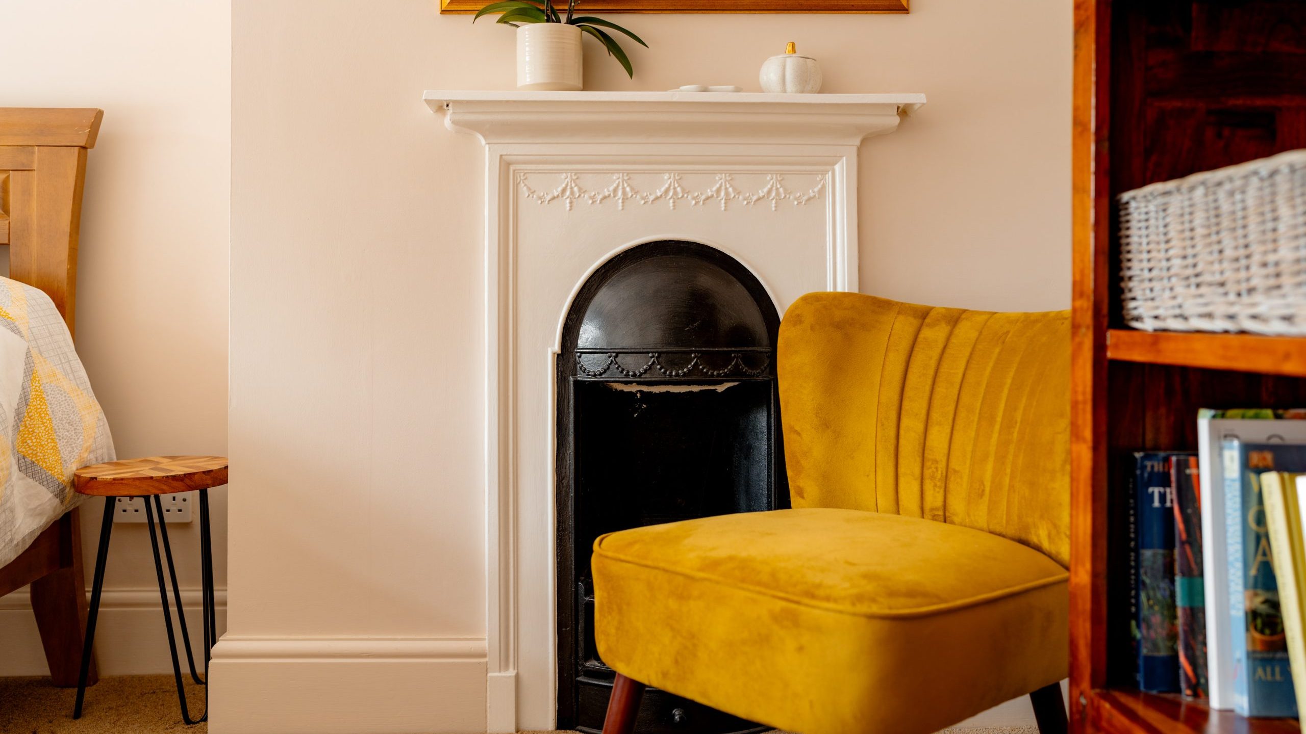 A cozy room featuring a mustard yellow accent chair in front of a white mantel with an ornamental design. On the mantel are a small white vase with a plant and a decorative item. Partial views of a wooden bed with a yellow blanket and a bookshelf are seen in the background.