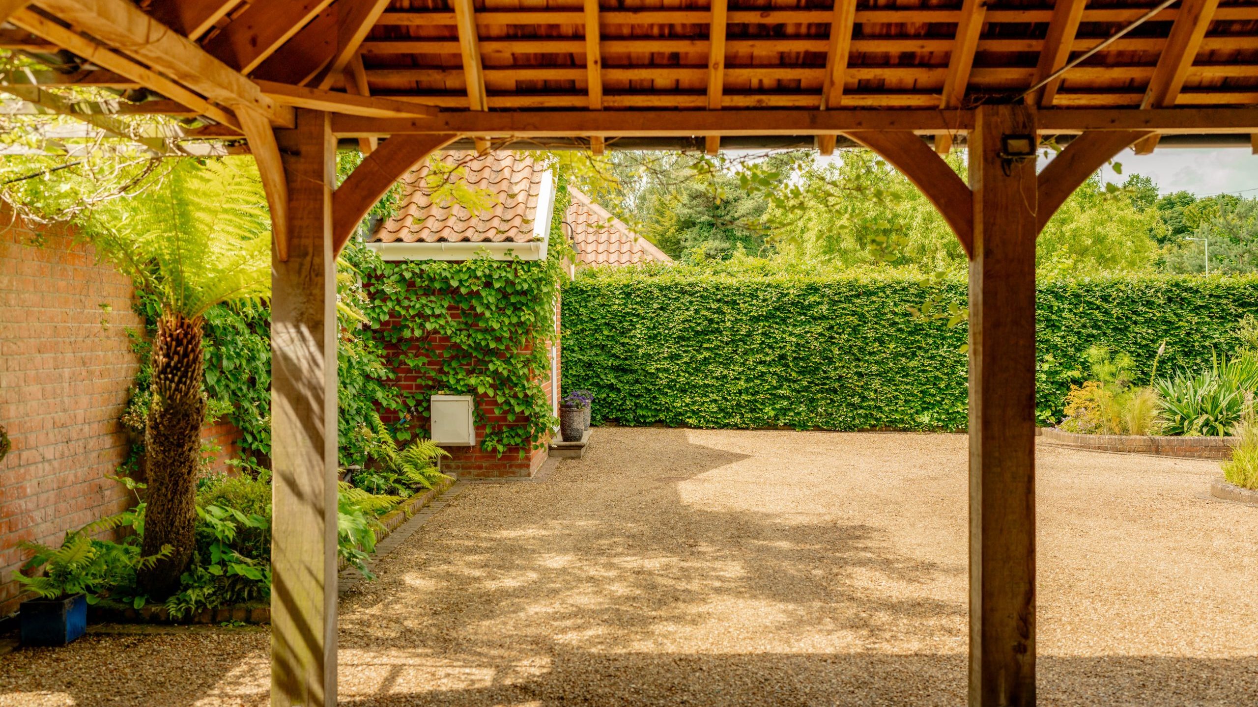 A wooden pergola with a tiled roof stands in the foreground, overlooking a sunny courtyard with gravel ground. In the background, a brick wall and a house covered in lush green ivy are visible. The courtyard is surrounded by a tall, well-maintained hedge.