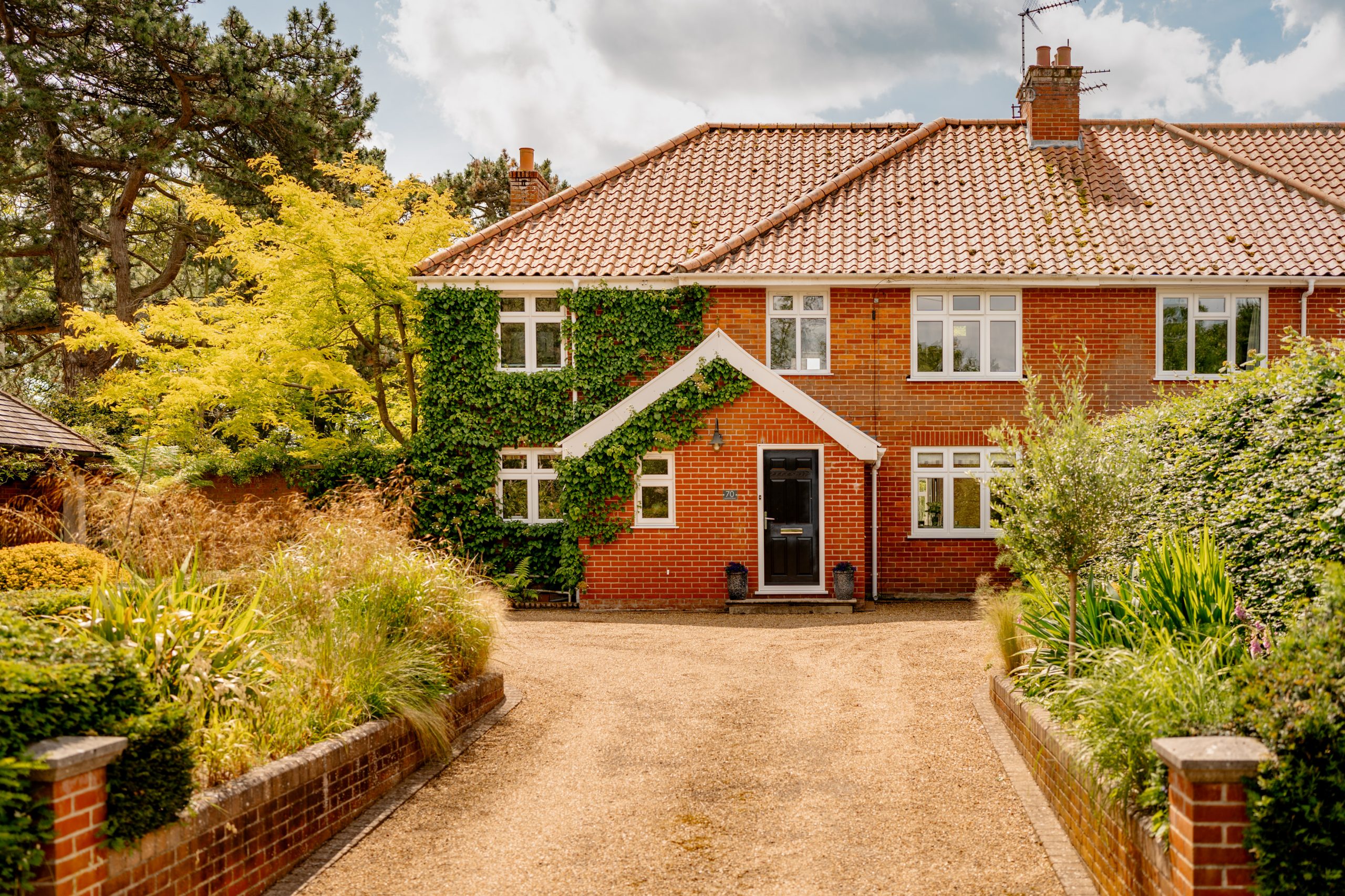 A charming two-story, red-brick house with a gabled roof and ivy climbing the front façade. It has white-framed windows and a black front door. The home is surrounded by lush greenery and colorful bushes, and a gravel driveway leads up to the entrance.