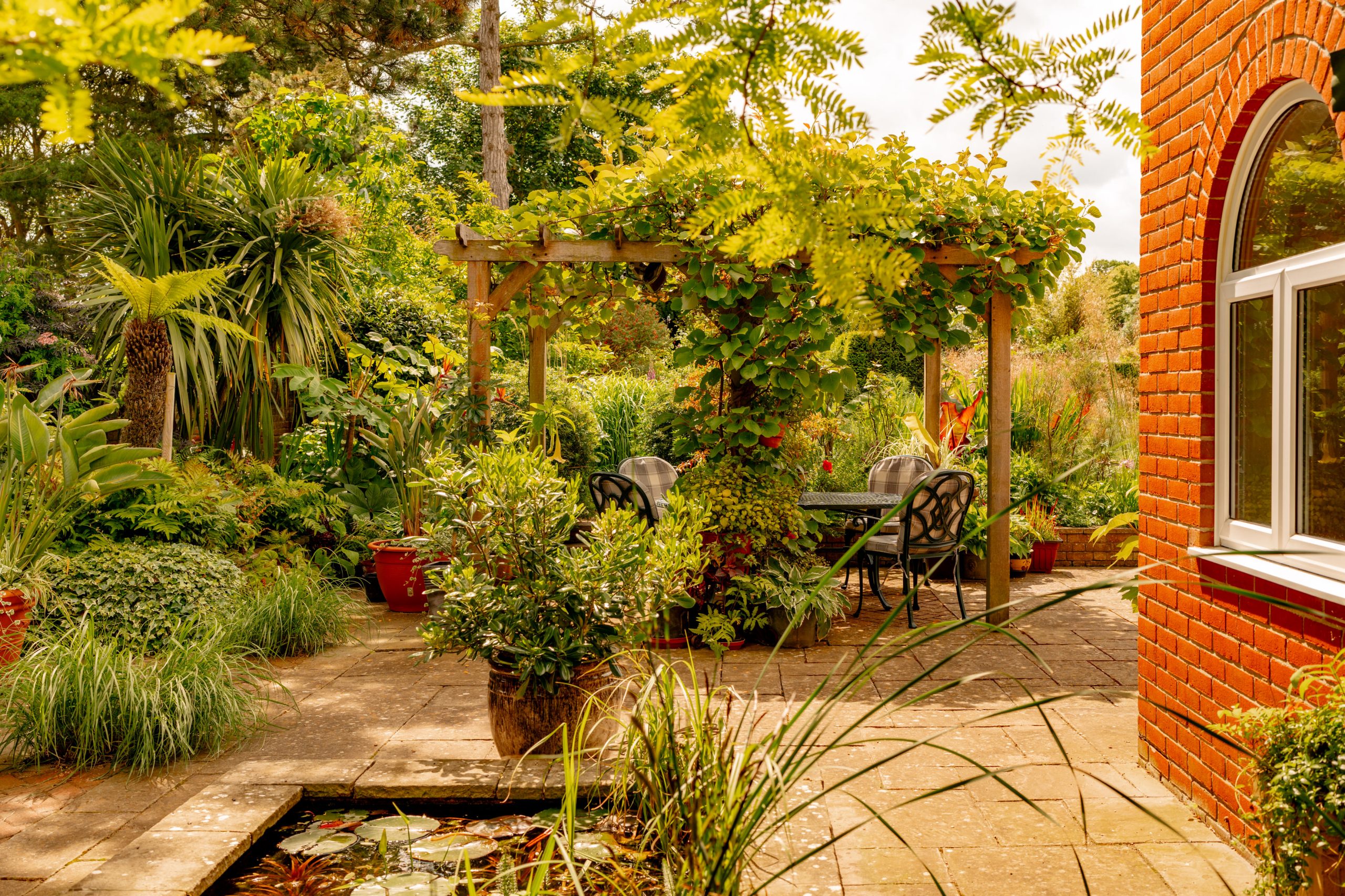 A lush garden scene with a vine-covered pergola shading a patio table and chairs. The patio is surrounded by various green plants and trees. A small pond is visible in the foreground, and a red brick house with a large arched window is on the right.