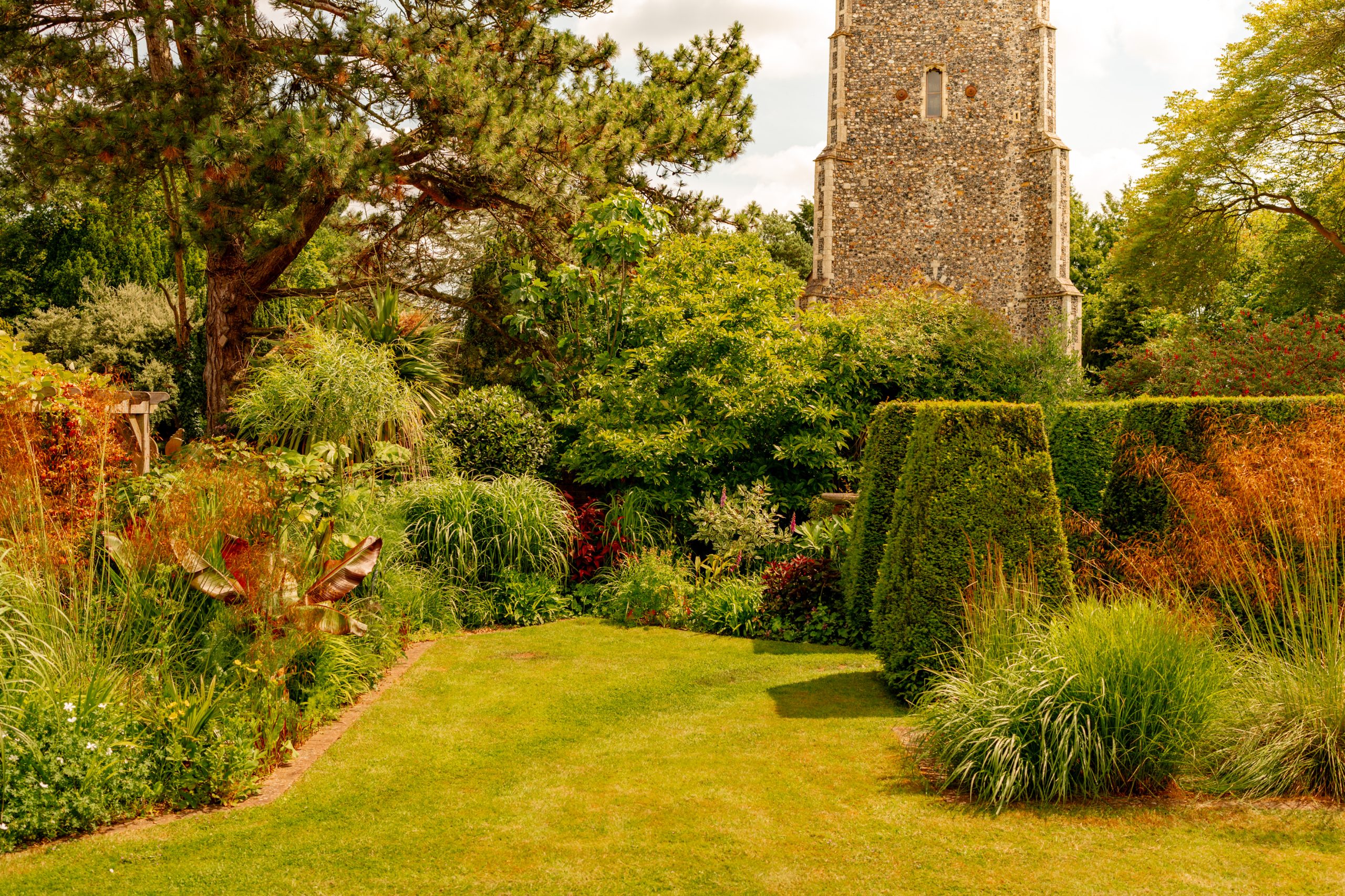 A lush garden featuring varied greenery, vibrant plants, and neatly trimmed hedges. In the background, a tall stone tower partially obscured by trees and shrubs stands against a blue sky dotted with clouds. The scene is bright and serene.