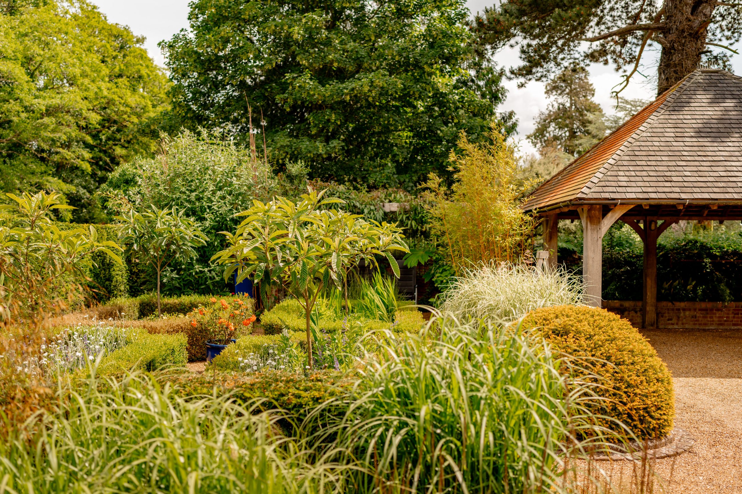 A lush garden featuring manicured bushes, tall grasses, and a variety of trees under a bright sky. To the right, there is a wooden gazebo with a shingled roof, surrounded by well-maintained plants and vibrant greenery.