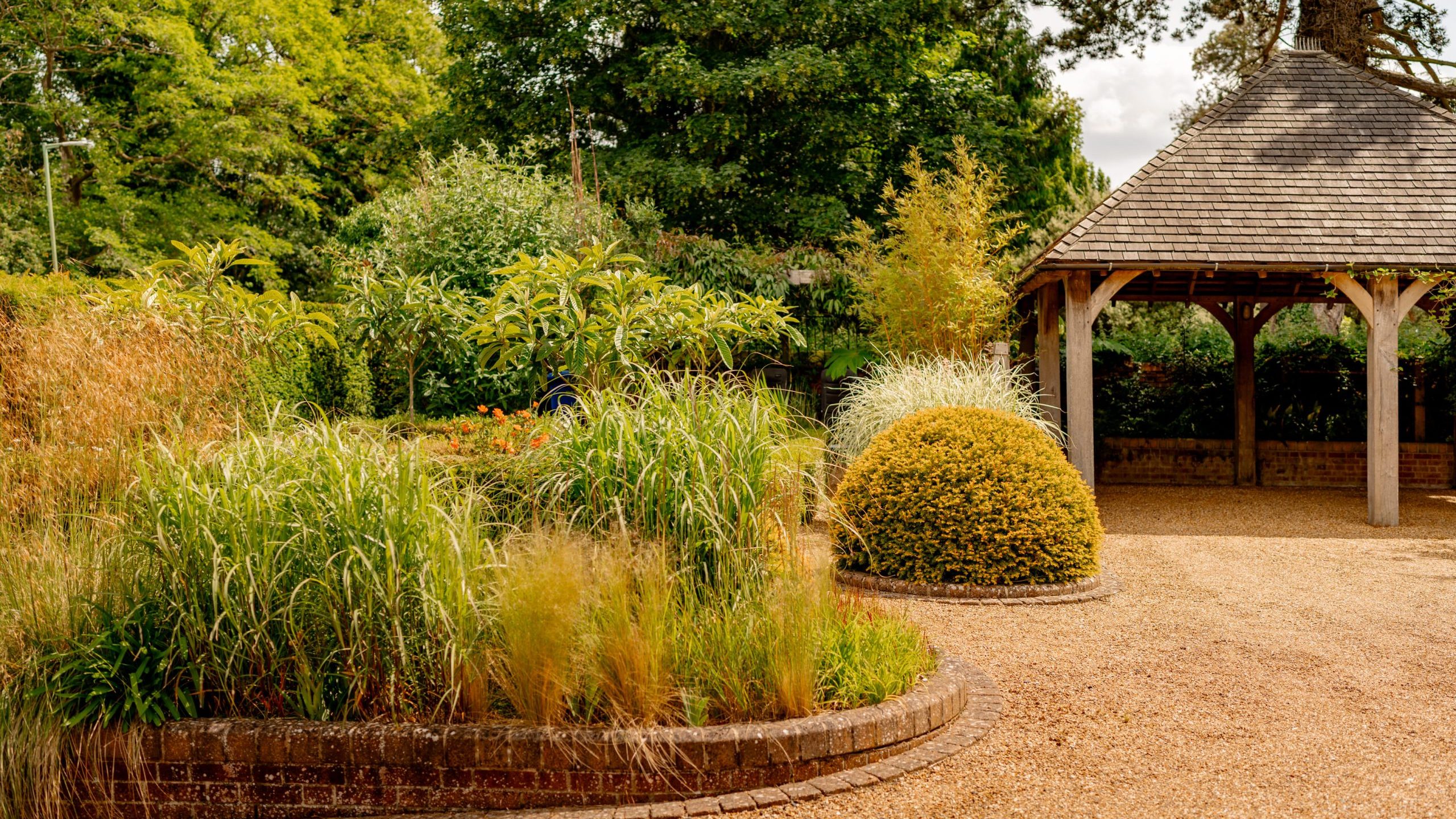 A well-maintained garden features a variety of green plants and tall grasses bordered by a low brick wall. A gravel path leads to a rustic gazebo with a shingle roof. Trees and dense foliage create a serene backdrop on a sunny day.