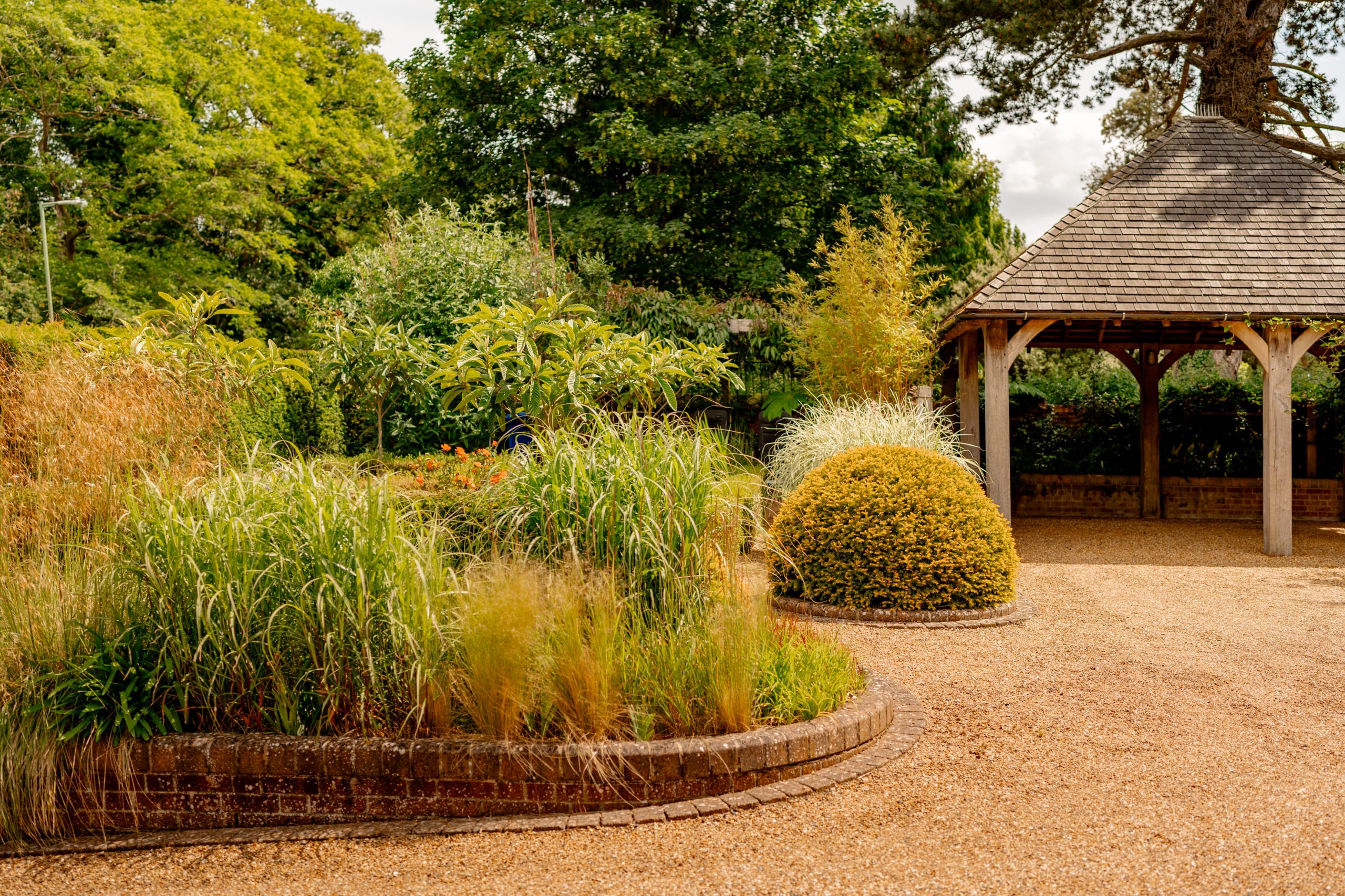 A well-maintained garden features a variety of green plants and tall grasses bordered by a low brick wall. A gravel path leads to a rustic gazebo with a shingle roof. Trees and dense foliage create a serene backdrop on a sunny day.