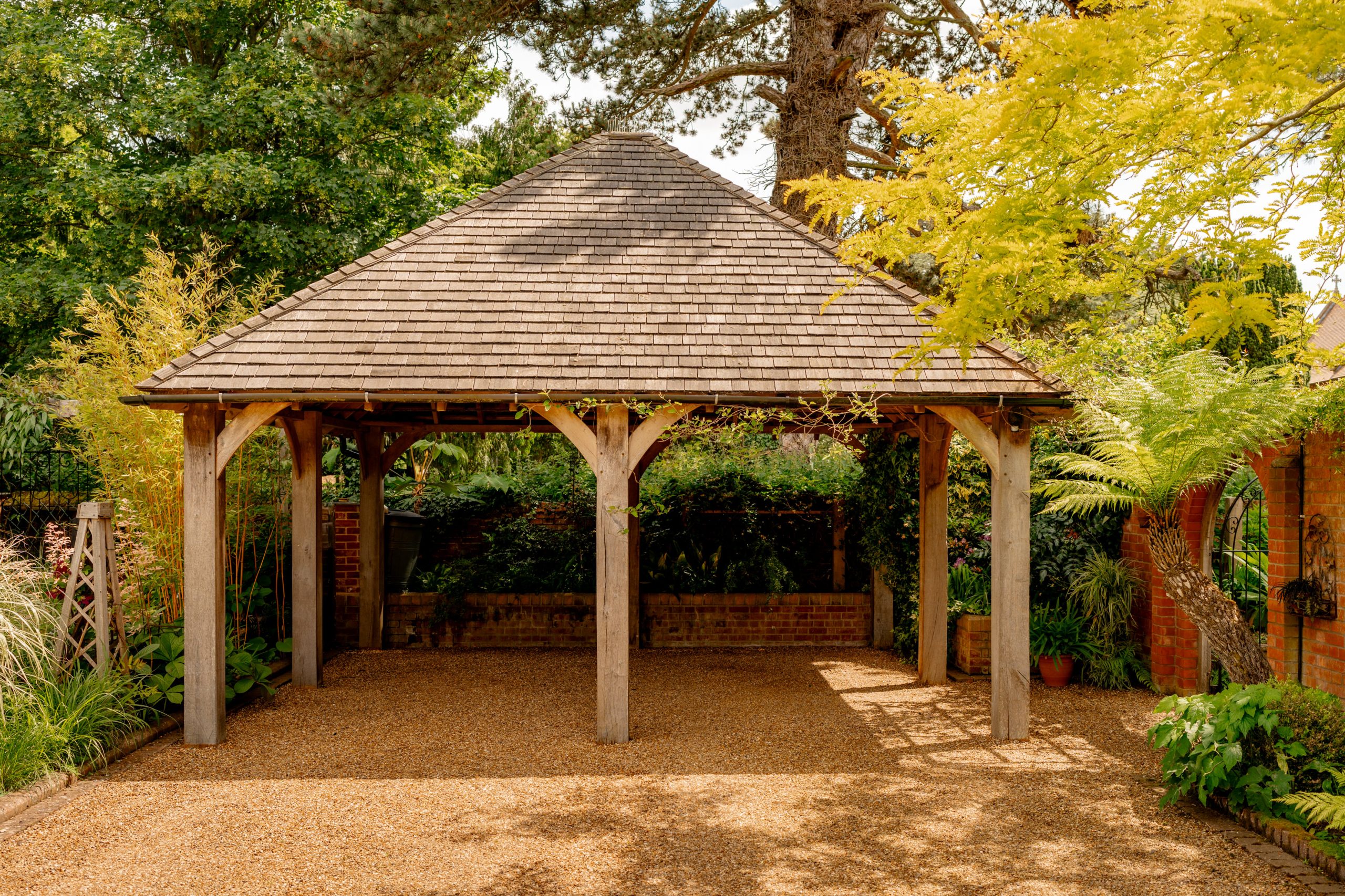 A wooden carport with a shingled roof stands in a shaded garden area, surrounded by lush greenery, including trees, shrubs, and potted plants. The sunlight filters through the leaves, casting dappled shadows on the gravel ground beneath the structure.