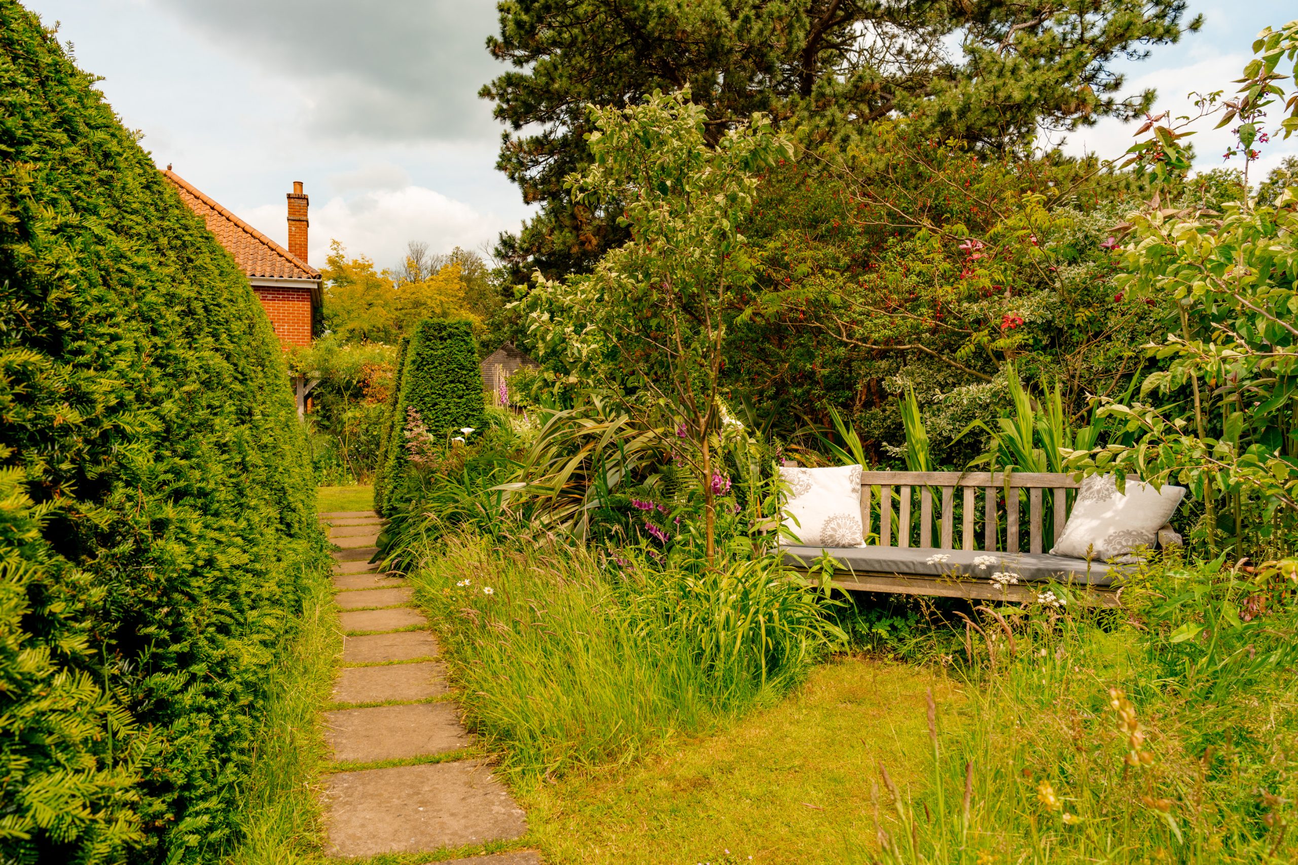 A lush garden with a stone path leading to a red-roofed house. On the right, a wooden bench with two white pillows sits among flowering plants and tall greenery. The scene is peaceful with a mix of sunlight and shade from the trees.