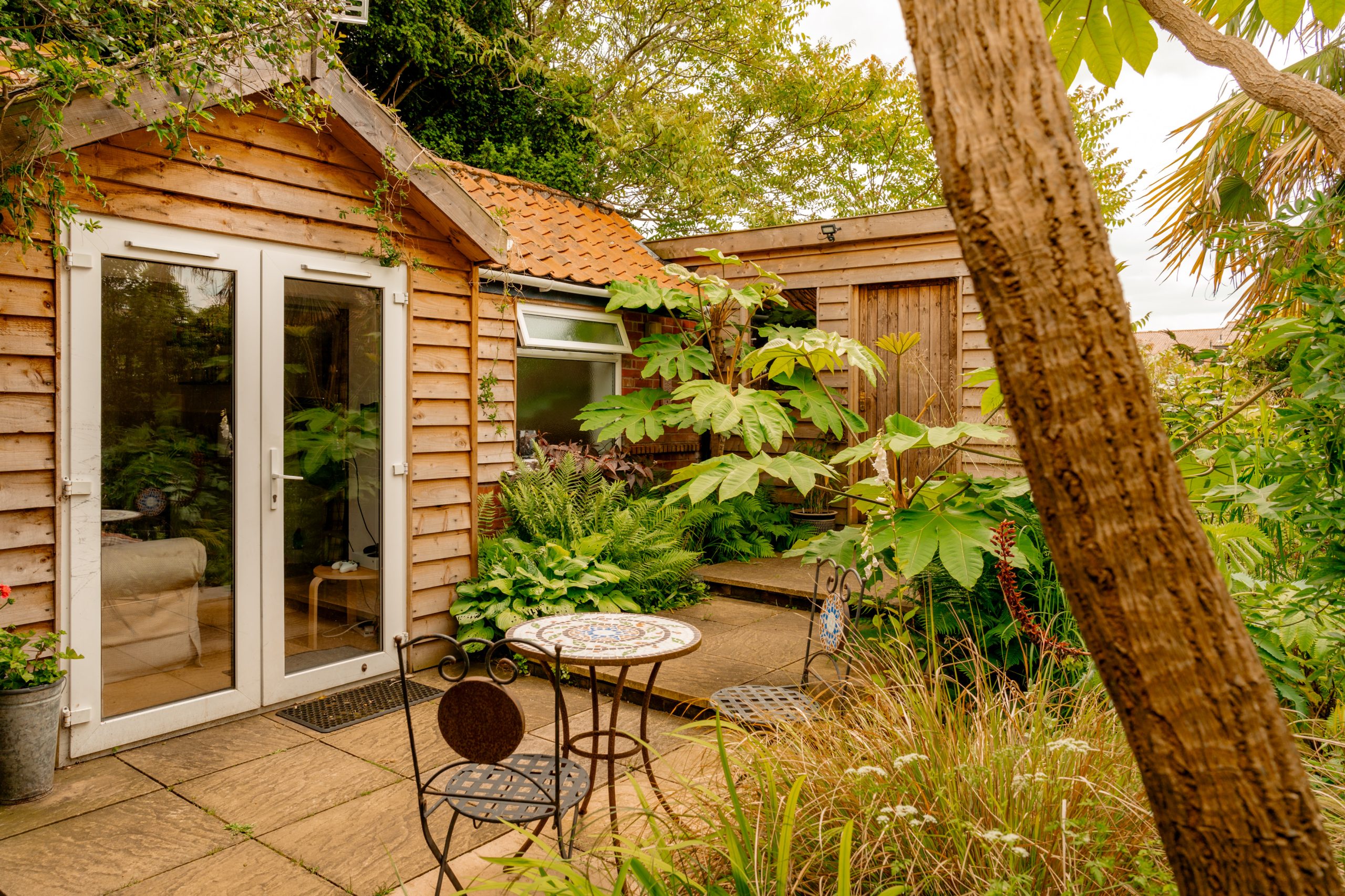 A charming wooden garden shed with a tiled roof stands amid lush greenery. It features glass double doors, a small round table with two chairs on a stone patio, and an abundance of plants and trees surrounding the structure, creating a serene atmosphere.