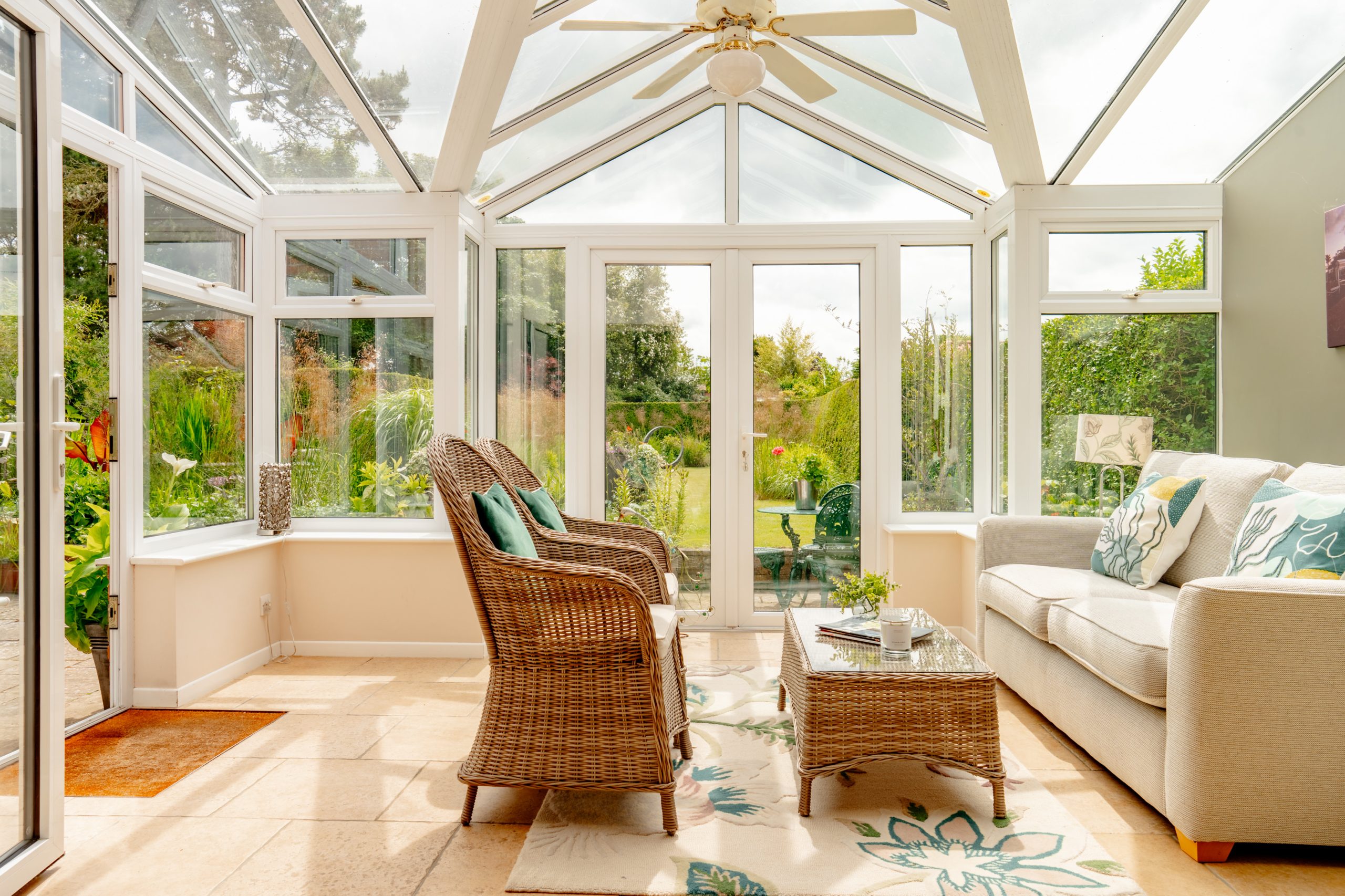 A bright conservatory with glass walls and ceiling features wicker furniture, including two chairs with cushions, a glass-top coffee table, and a beige sofa with decorative pillows. A garden with lush greenery is visible outside through the glass doors.
