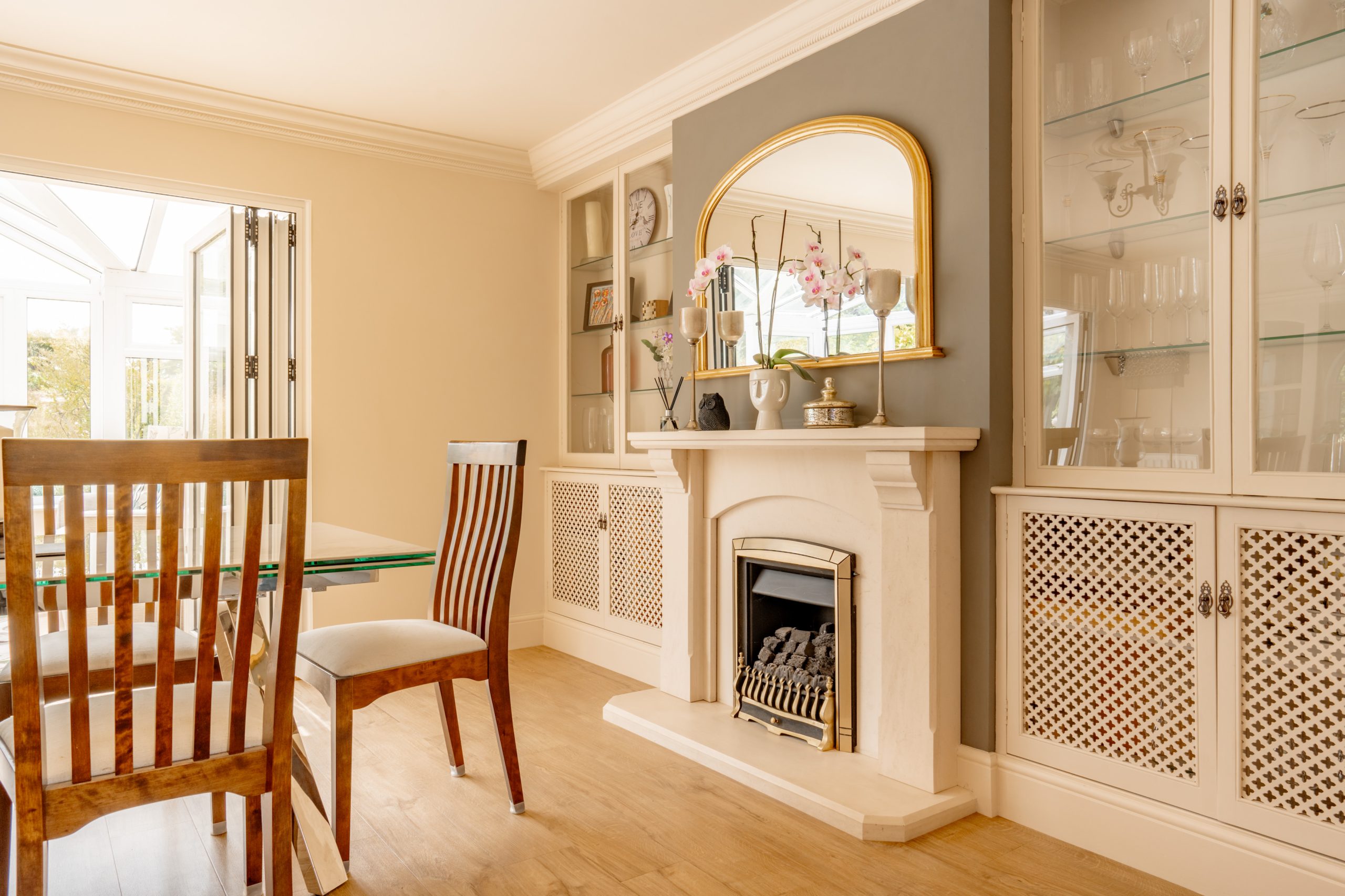 A cozy dining room features a wooden table and chairs, a fireplace with an ornate gold-framed mirror above it, and built-in shelves and cabinetry. A glass door opens out to a sunlit patio. The room is adorned with decorative items and has a warm, inviting ambiance.