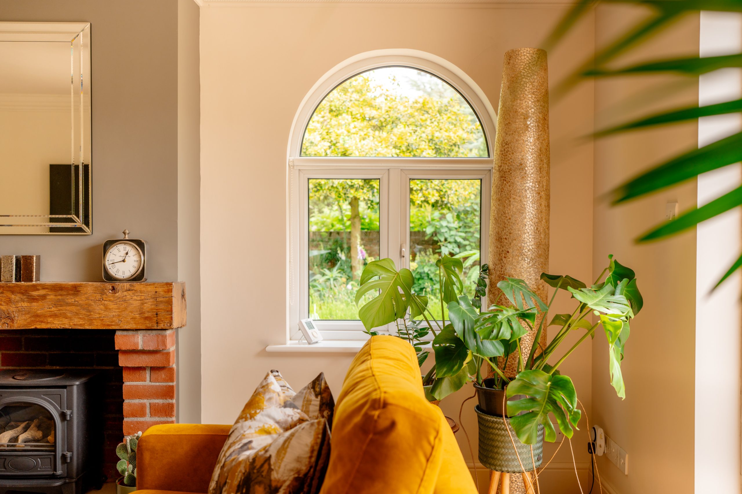 A cozy living room featuring a vibrant yellow sofa adorned with patterned cushions. A large potted plant sits by an arched window with views of a green garden. A brick fireplace holds a clock and mirror, while a tall, textured vase stands nearby.