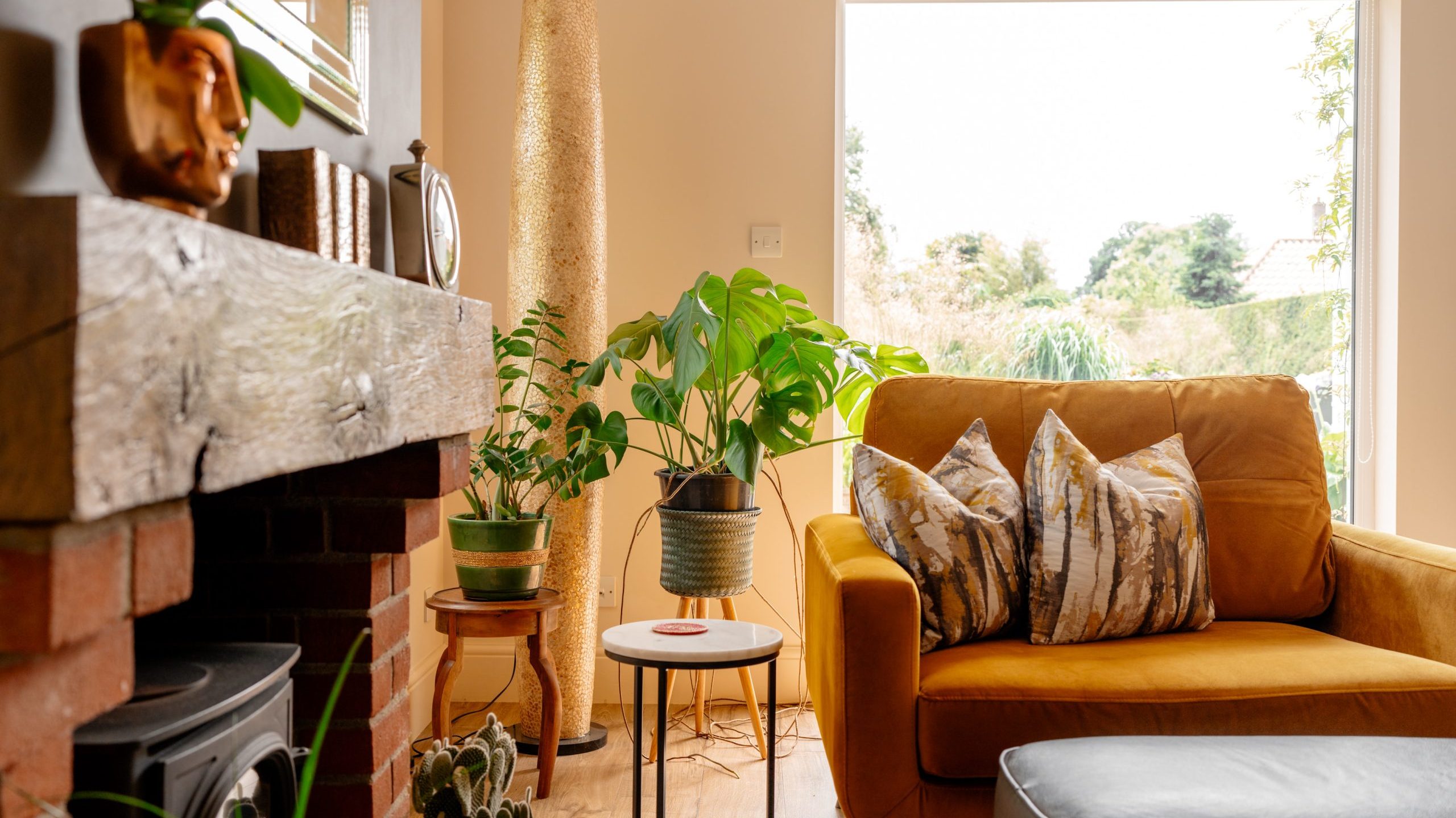 A cozy living room with a mustard yellow armchair adorned with patterned cushions. Surrounding the chair are various indoor plants in pots. In the background, a large window lets in natural light, highlighting the room’s soft earthy tones and wooden flooring.