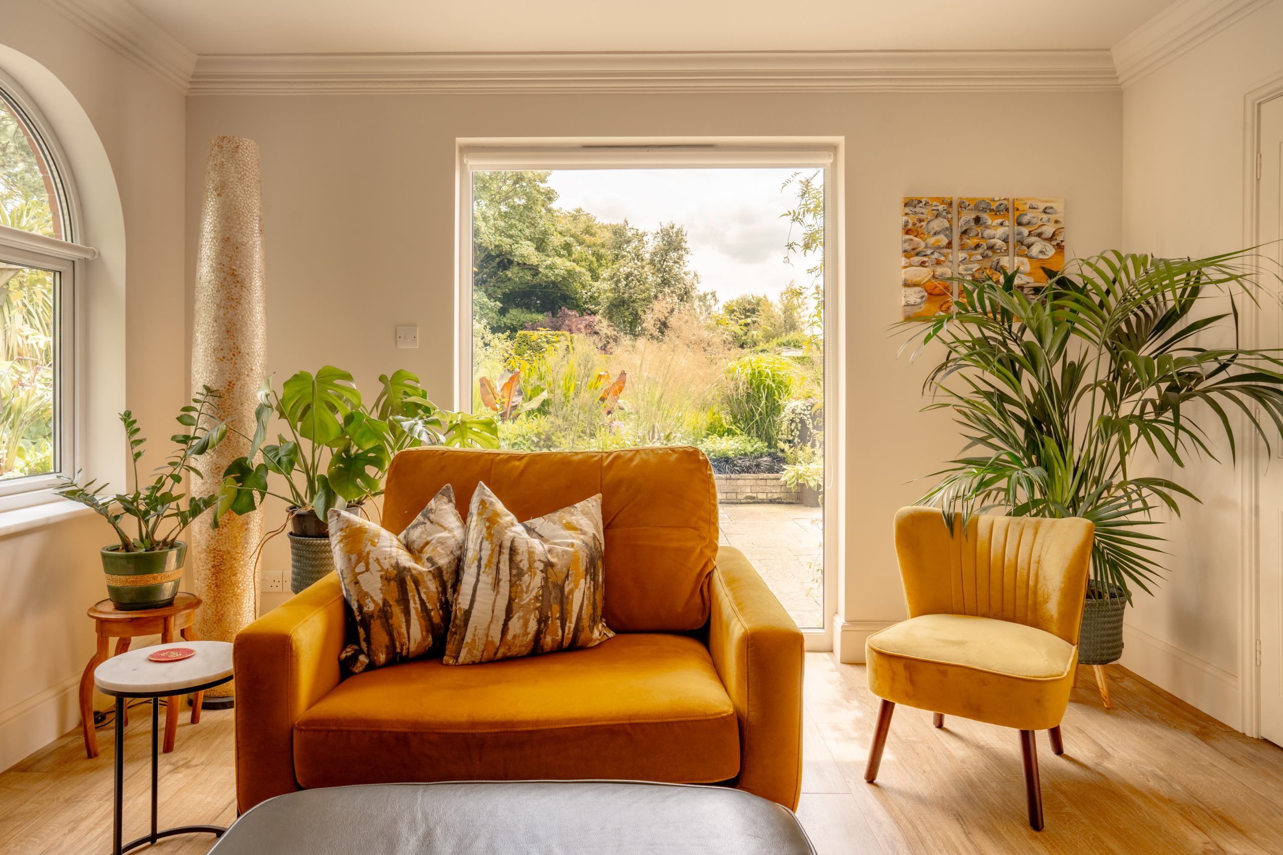 A cozy living room with a large yellow armchair and a matching yellow chair. The room has several potted plants and a large window offering a view of a lush garden outside. Natural light floods the space, enhancing the warm, inviting atmosphere.
