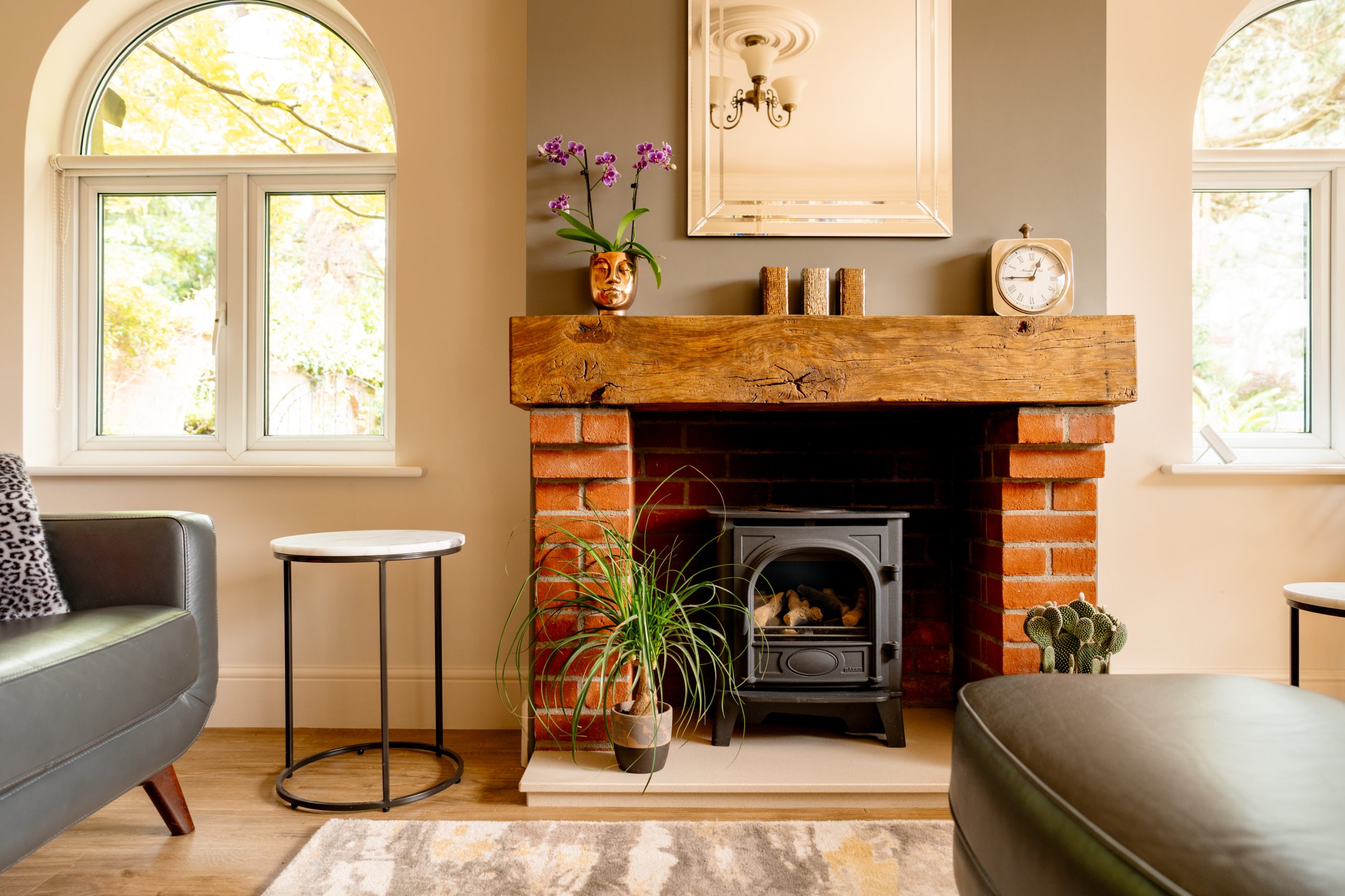 A cozy living room features a brick and wood-framed fireplace with a wood-burning stove. A wooden mantel holds a potted orchid, candles, and a clock. Flanked by arched windows, the room includes a round side table, houseplants, and part of a leather sofa and ottoman.