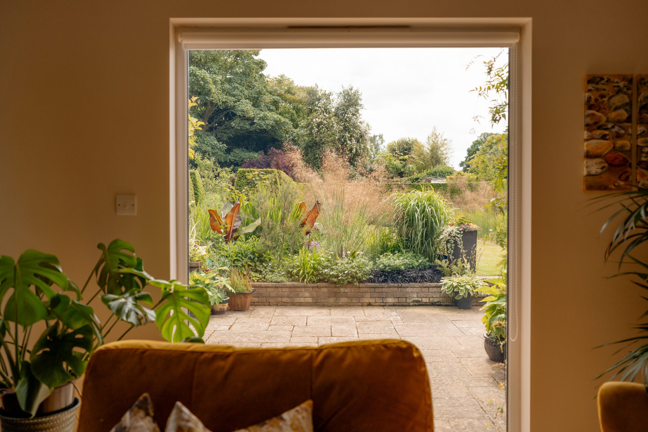 A view through a large open window showing a lush, green garden with various plants and trees. The patio has potted plants and is framed by indoor plants on both sides. The foreground has a cozy couch with patterned pillows. The sky is overcast with soft light.