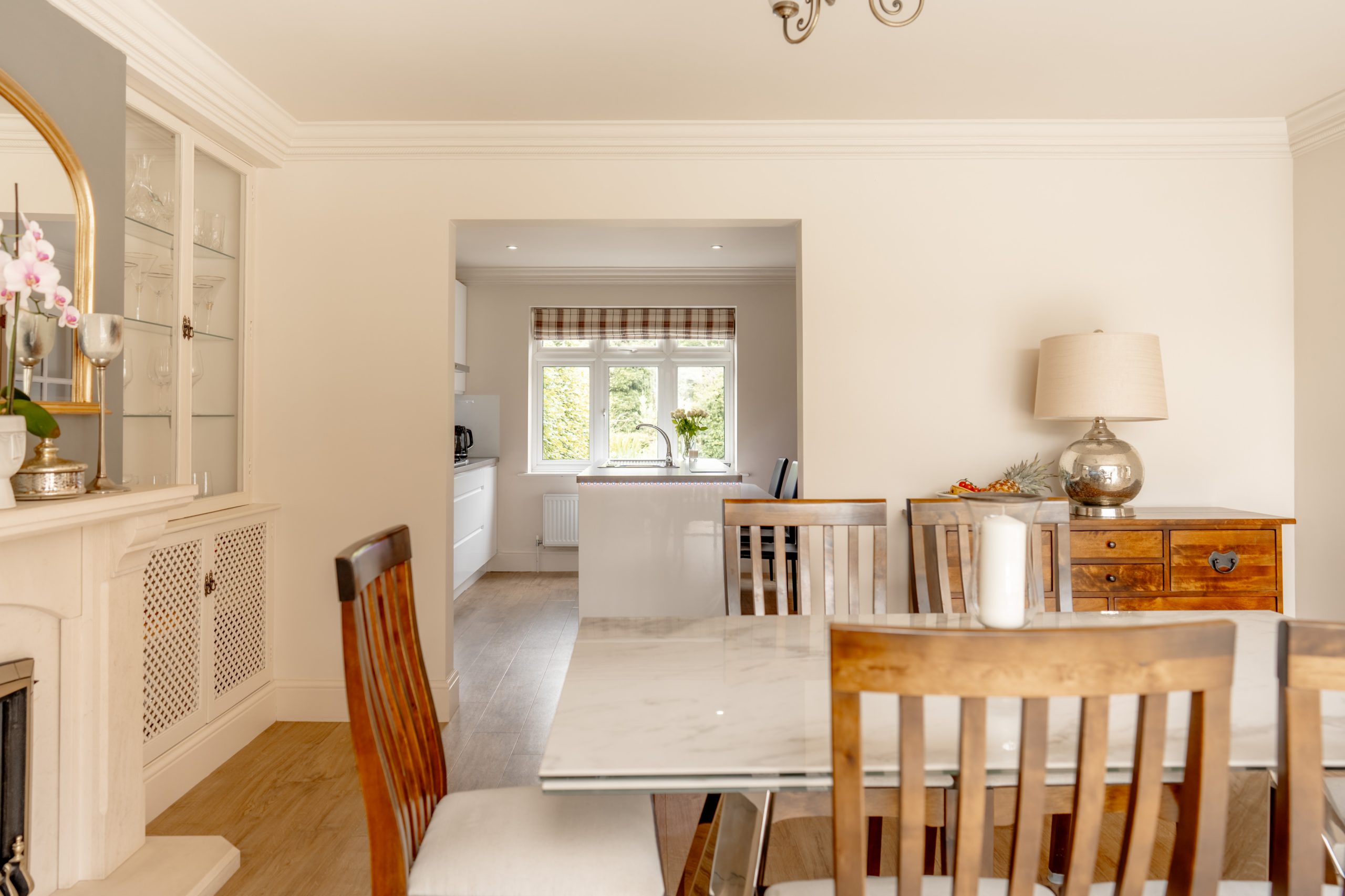 A cozy dining room with a marble table and wooden chairs. To the left is a fireplace with a decorative mirror and vase. At the back, an entryway leads to a bright kitchen with a window featuring a striped valance. A wooden sideboard with a lamp stands on the right.