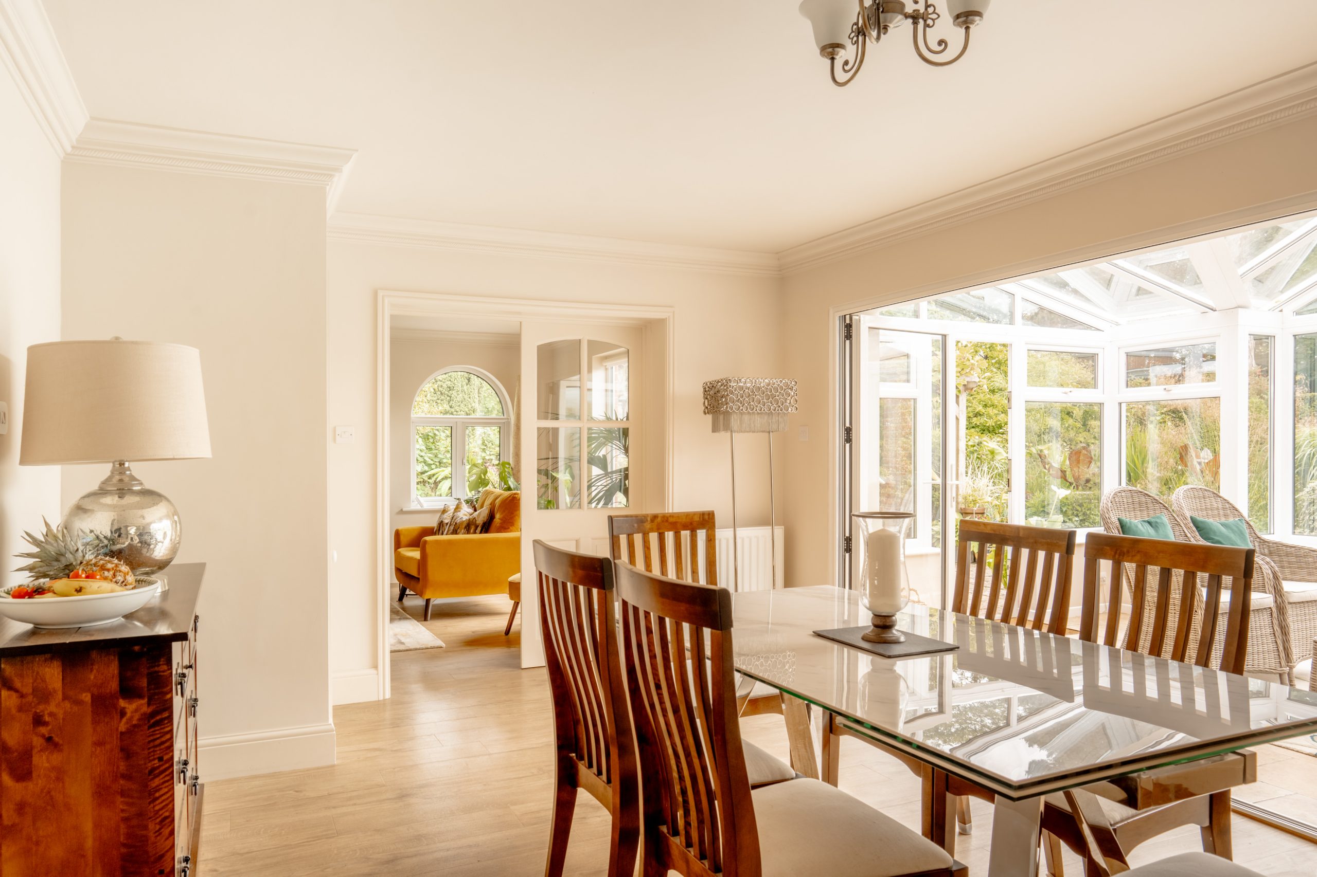 A bright, airy dining room features a glass-top table with wooden chairs. A fruit bowl sits on a sideboard with a lamp nearby. Large windows and French doors open to a sunroom with wicker chairs, allowing natural light to flood the space. A cozy seating area is visible.