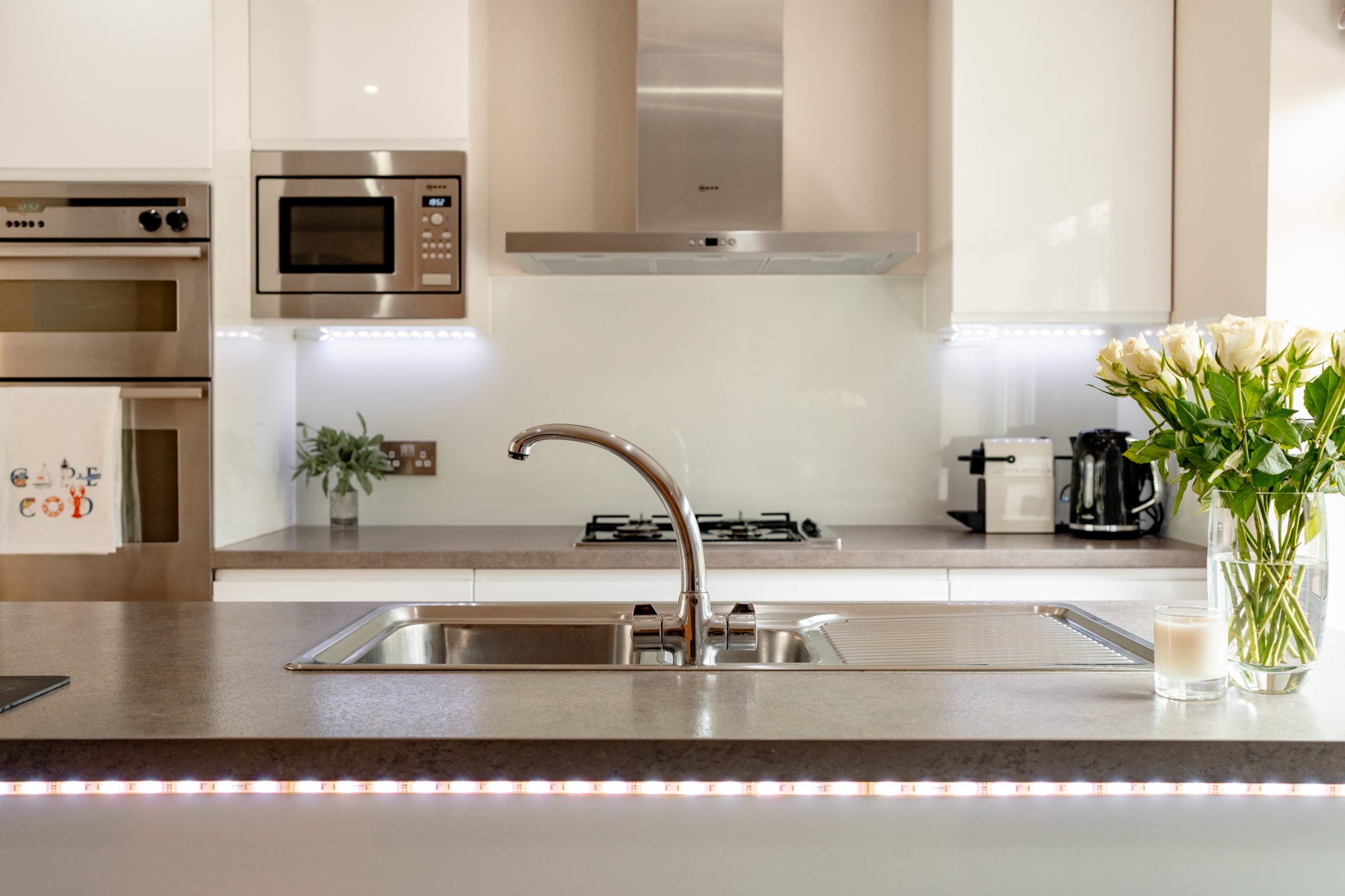 A modern kitchen featuring a central island with a sink and stainless steel faucet. Behind the island, there are stainless steel appliances, including a microwave and an oven. A bouquet of white roses sits on the counter, with a coffee maker and a candle nearby.