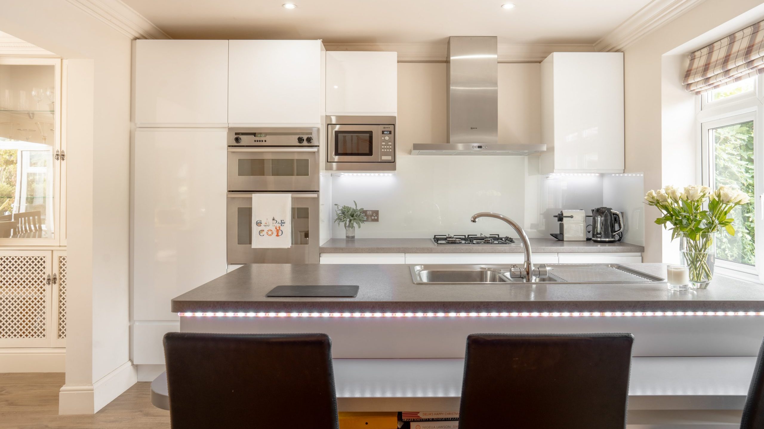 Modern kitchen with white cabinets, stainless steel oven, microwave, and range hood. An island features a built-in sink, mixer tap, and induction cooktop, with three black barstools. A vase of white flowers is on the counter near a window with striped blinds.