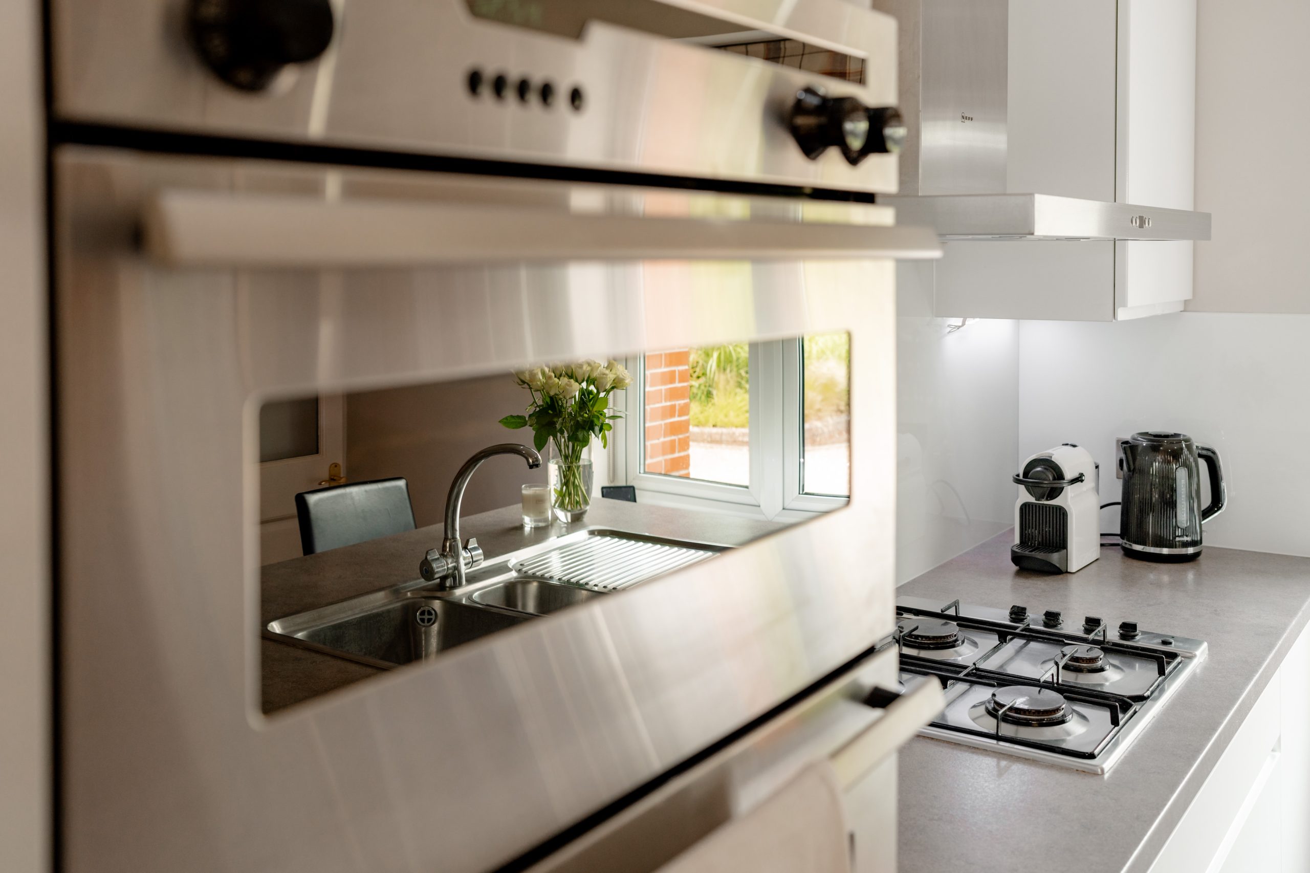 Modern kitchen with stainless steel appliances including a built-in oven, gas stovetop, and a kettle on the countertop. A sink is beneath a window, with sunlight illuminating a vase of flowers on the windowsill. White cabinetry provides a clean look.