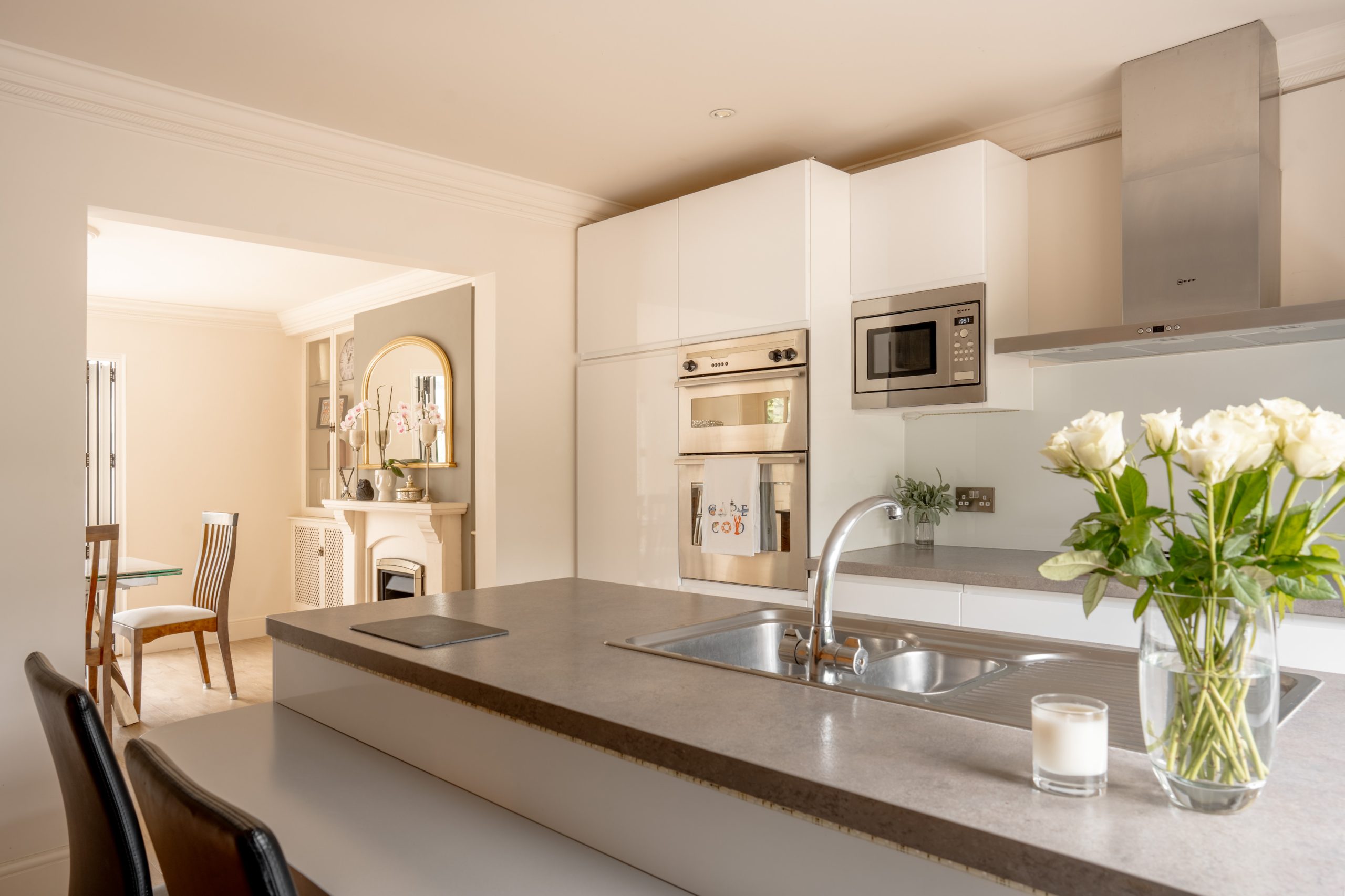 Modern kitchen with a gray countertop and white cabinetry. The center island has a sink and two black stools. Built-in appliances include a microwave and oven. A vase with white roses and a candle decorate the counter. The dining area is visible in the background.