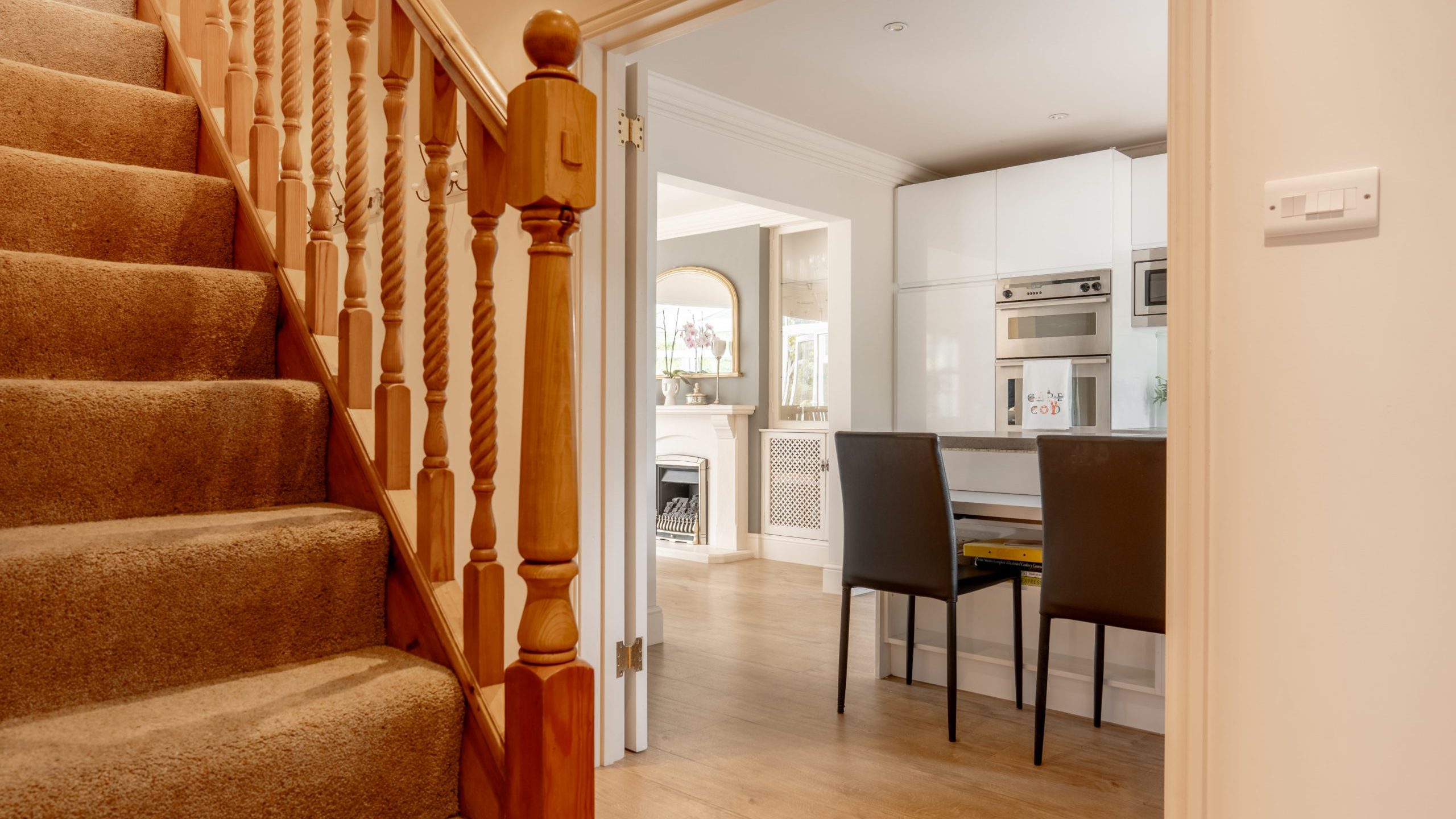 A cozy home interior featuring a carpeted staircase on the left with wooden railings, leading to an open kitchen and dining area. The kitchen has white cabinetry, built-in appliances, a small dining table, and two dark-colored chairs. The space is warmly lit.