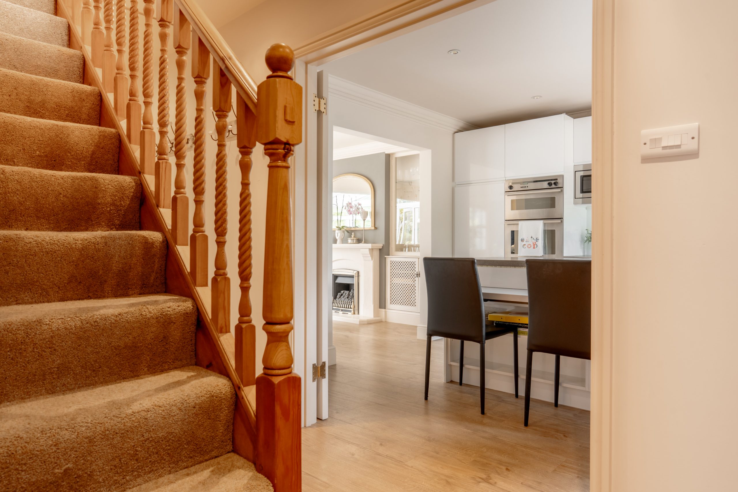 A cozy home interior featuring a carpeted staircase on the left with wooden railings, leading to an open kitchen and dining area. The kitchen has white cabinetry, built-in appliances, a small dining table, and two dark-colored chairs. The space is warmly lit.