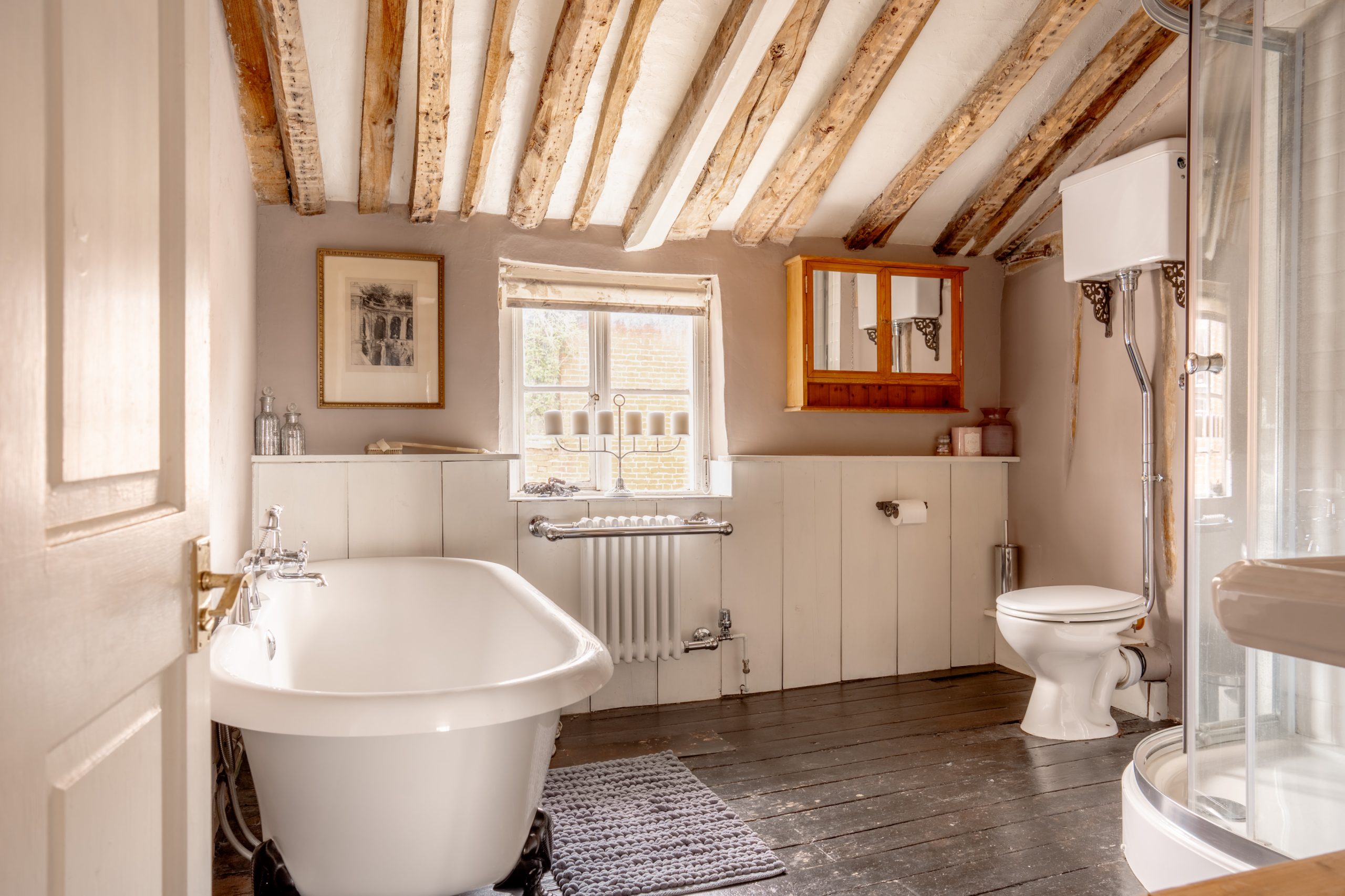 A rustic bathroom with exposed wooden beams on the ceiling, a white freestanding bathtub, a gray bath mat, a toilet, wall-mounted cabinets with a mirror, a radiator beneath a window, a framed picture, and a glass-enclosed shower in the corner.
