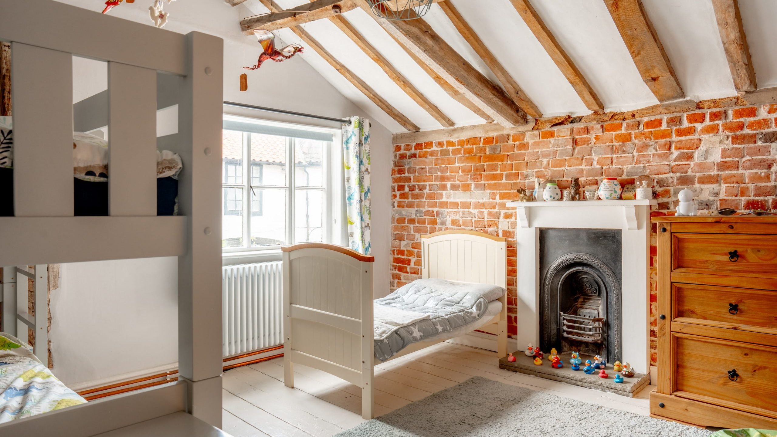 A cozy children's bedroom with exposed wooden beams and a brick accent wall. The room features a bunk bed, single bed, fireplace, wooden dresser, large window, and toys neatly arranged on the floor. Light streams in, creating a warm and inviting atmosphere.