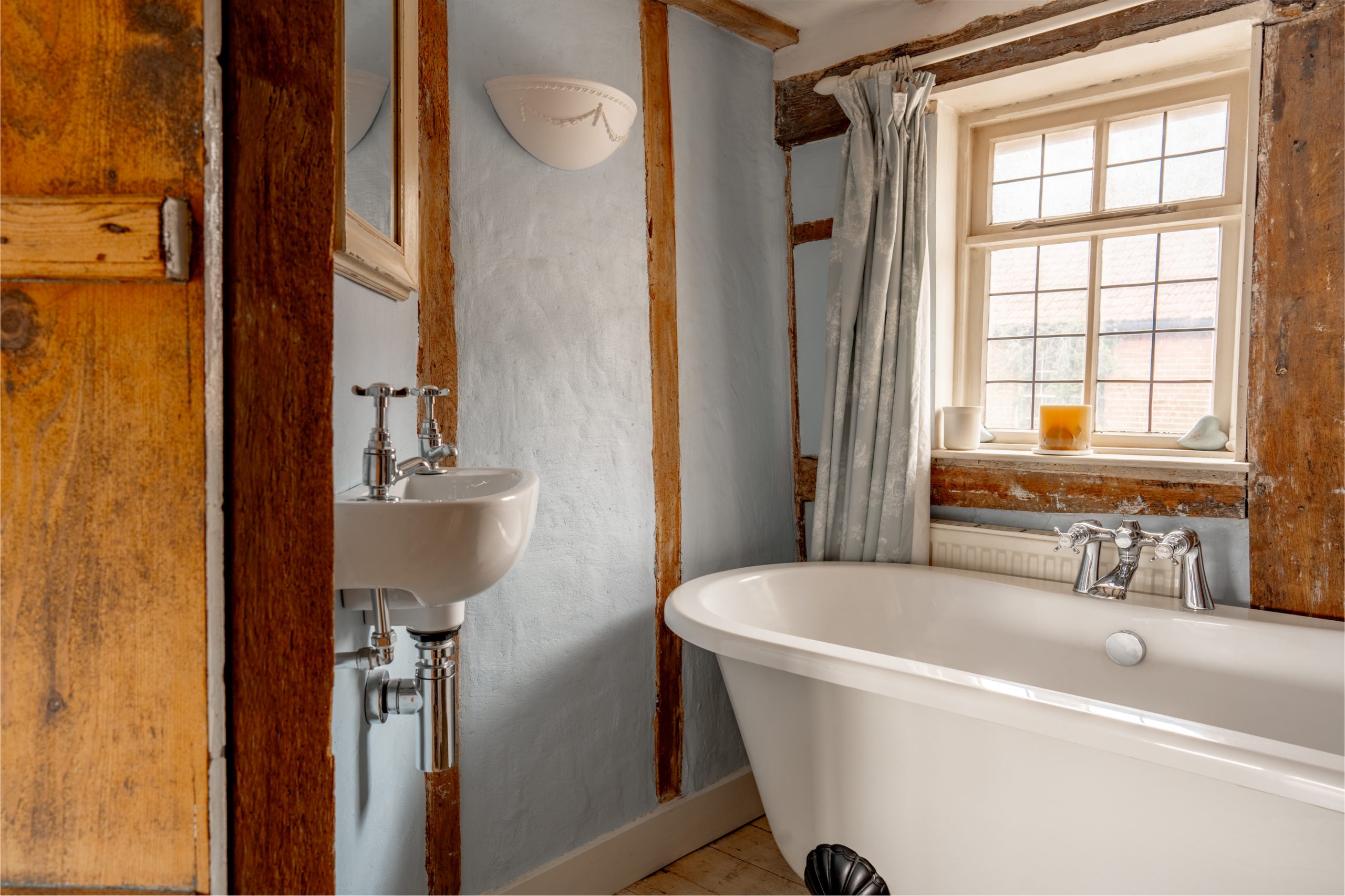 A rustic bathroom with wooden beams features a freestanding white bathtub, a small wall-mounted sink, and a pale blue curtain on the window. A candle sits on the windowsill, and a white wall sconce provides light. The walls are textured with a soft blue color.