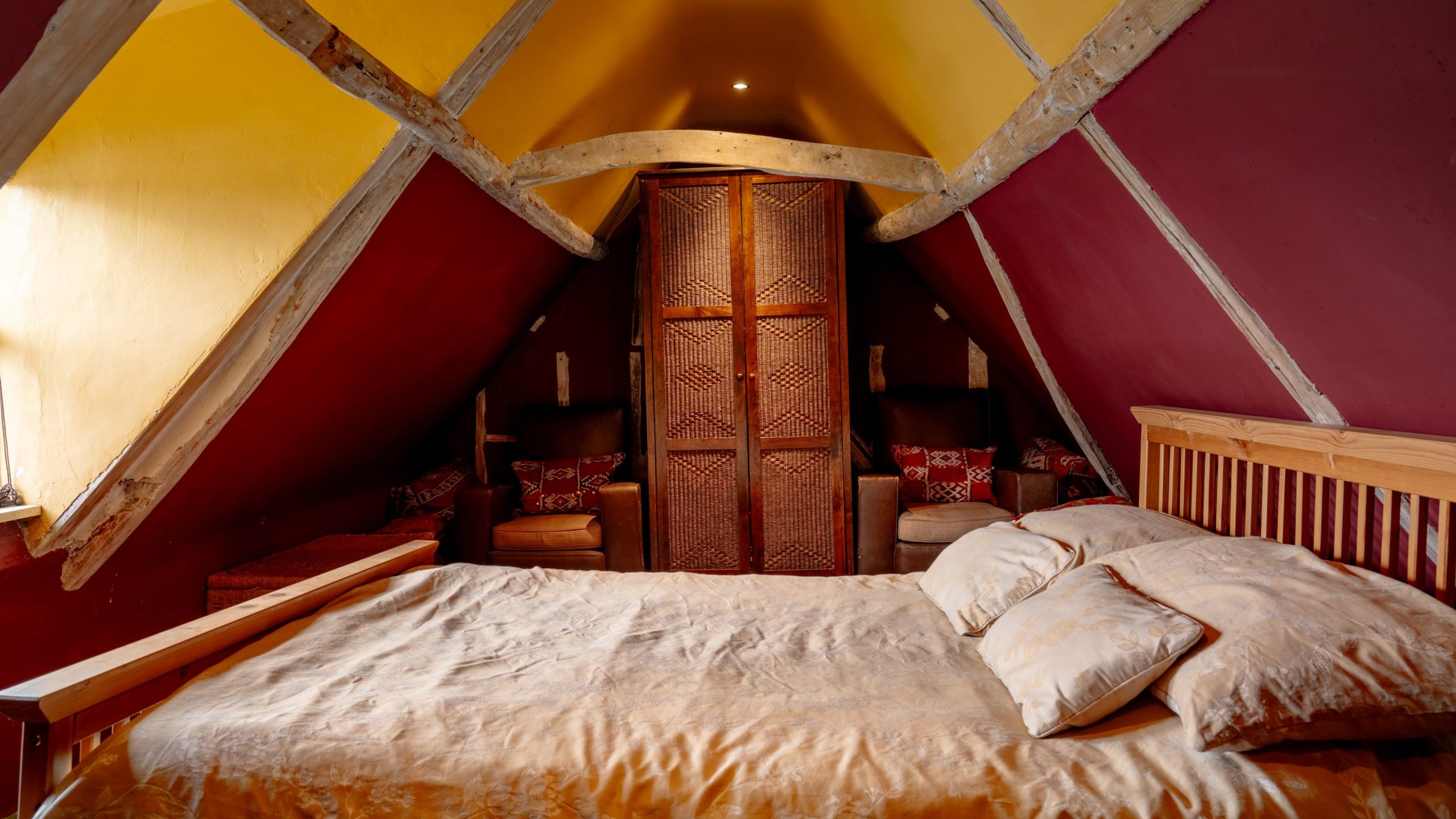 A cozy attic bedroom with a wooden bed covered in beige linens and pillows. The walls feature a vibrant mix of red and yellow, and the ceiling has exposed wooden beams. Two armchairs with decorative pillows flank a textured wooden wardrobe at the end of the room.