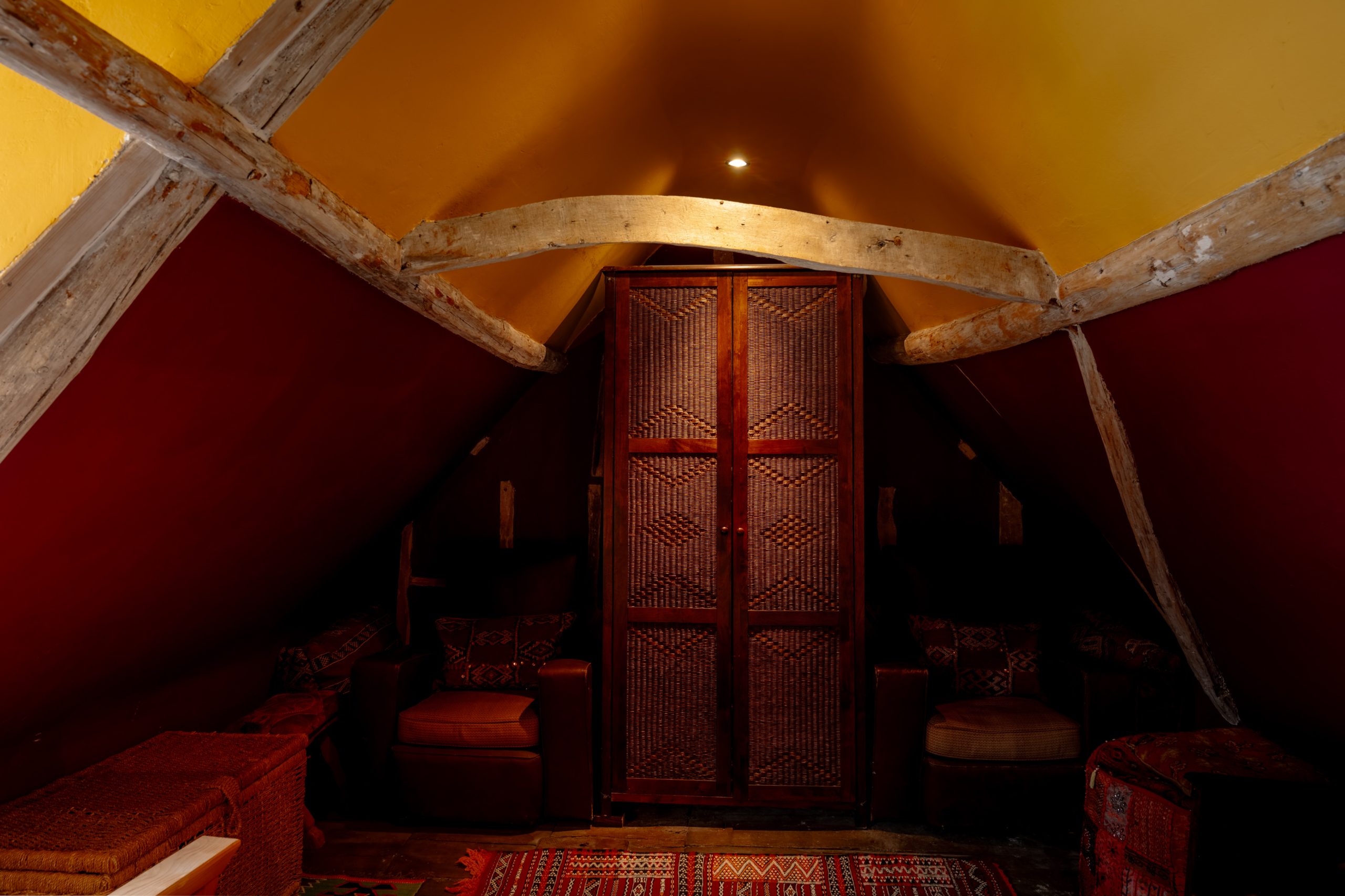 A cozy attic space with slanted ceilings, featuring a central wooden cabinet with intricate woven patterns. The room is dimly lit with warm yellow lighting, wooden beams, red walls, patterned rugs, and upholstered chairs on either side of the cabinet.