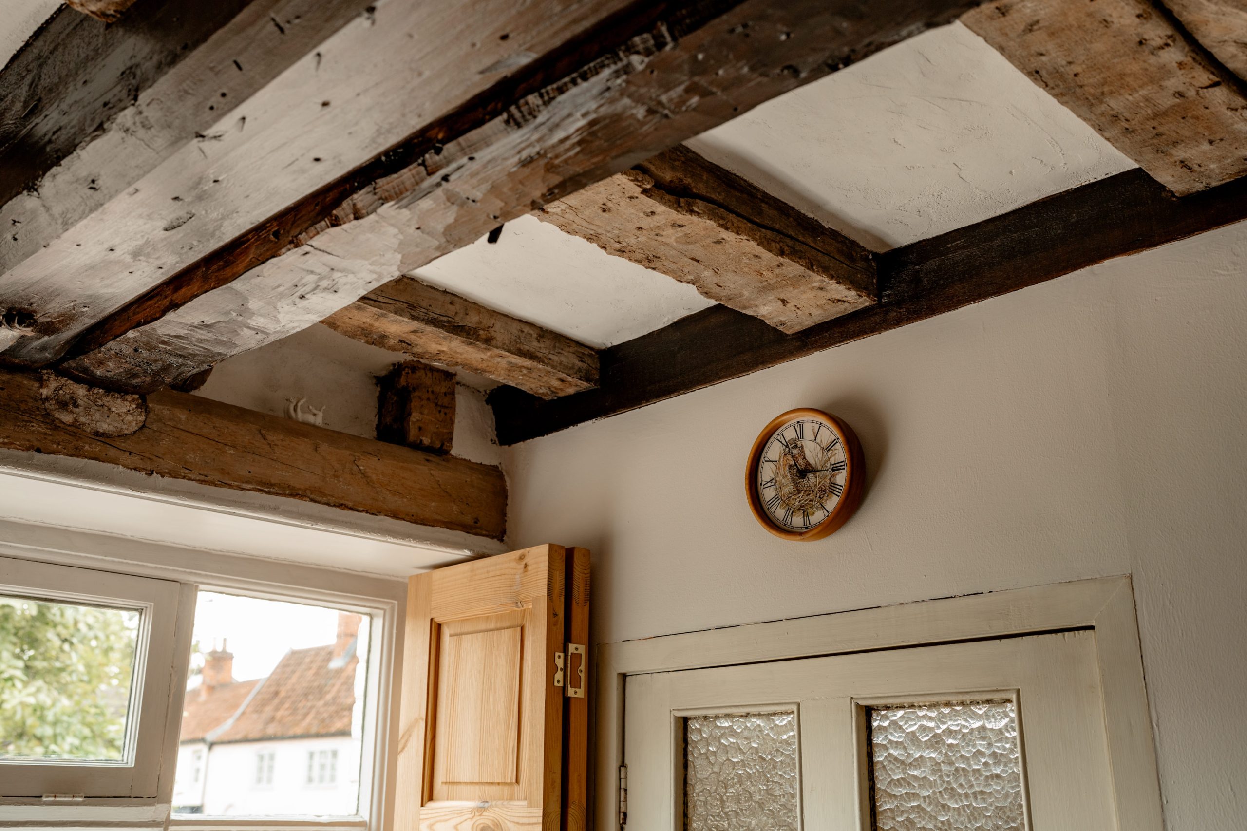 A rustic interior features exposed wooden beams on the ceiling, a round clock on the wall, a partially open wooden door, and a textured glass-paneled door. Outside the window, parts of a neighboring building and greenery are visible.
