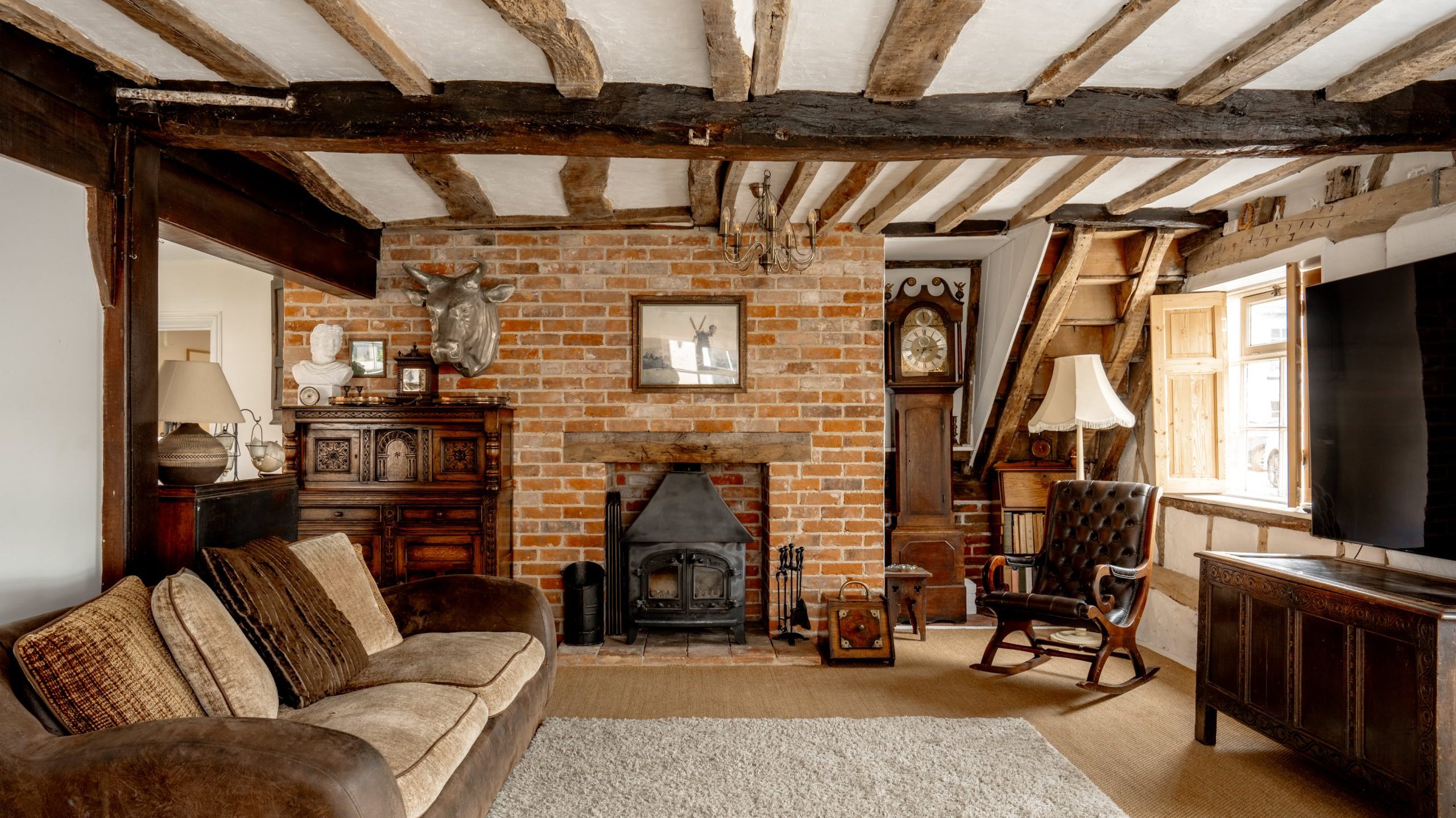 A cozy living room with exposed wooden beams on the ceiling, a brick fireplace, and a wood-burning stove. The room has a cushy brown sofa, a rocking chair, a wooden cabinet with a lamp, and a large TV. Paintings and a mounted deer head decorate the brick wall.
