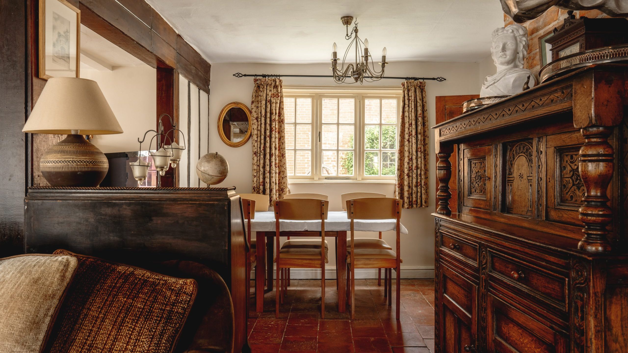 A cozy dining room with a wooden table and four chairs, lit by a window with patterned curtains. The room features vintage wooden furniture, including a cabinet and a sideboard with decorative items like a bust and a lamp. The floor is tiled in warm colors.