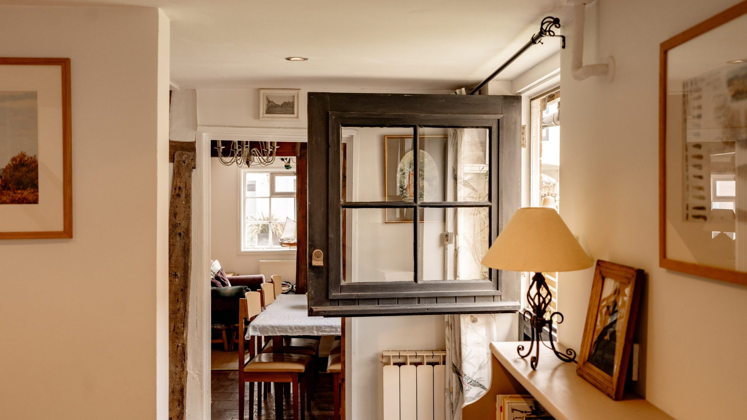 A cozy, rustic interior features a small dining area viewed through a vintage black-framed window mounted inside the house. The room is decorated with framed pictures, a table with chairs, a chandelier, and shelves holding books and a lamp. Natural light fills the space.