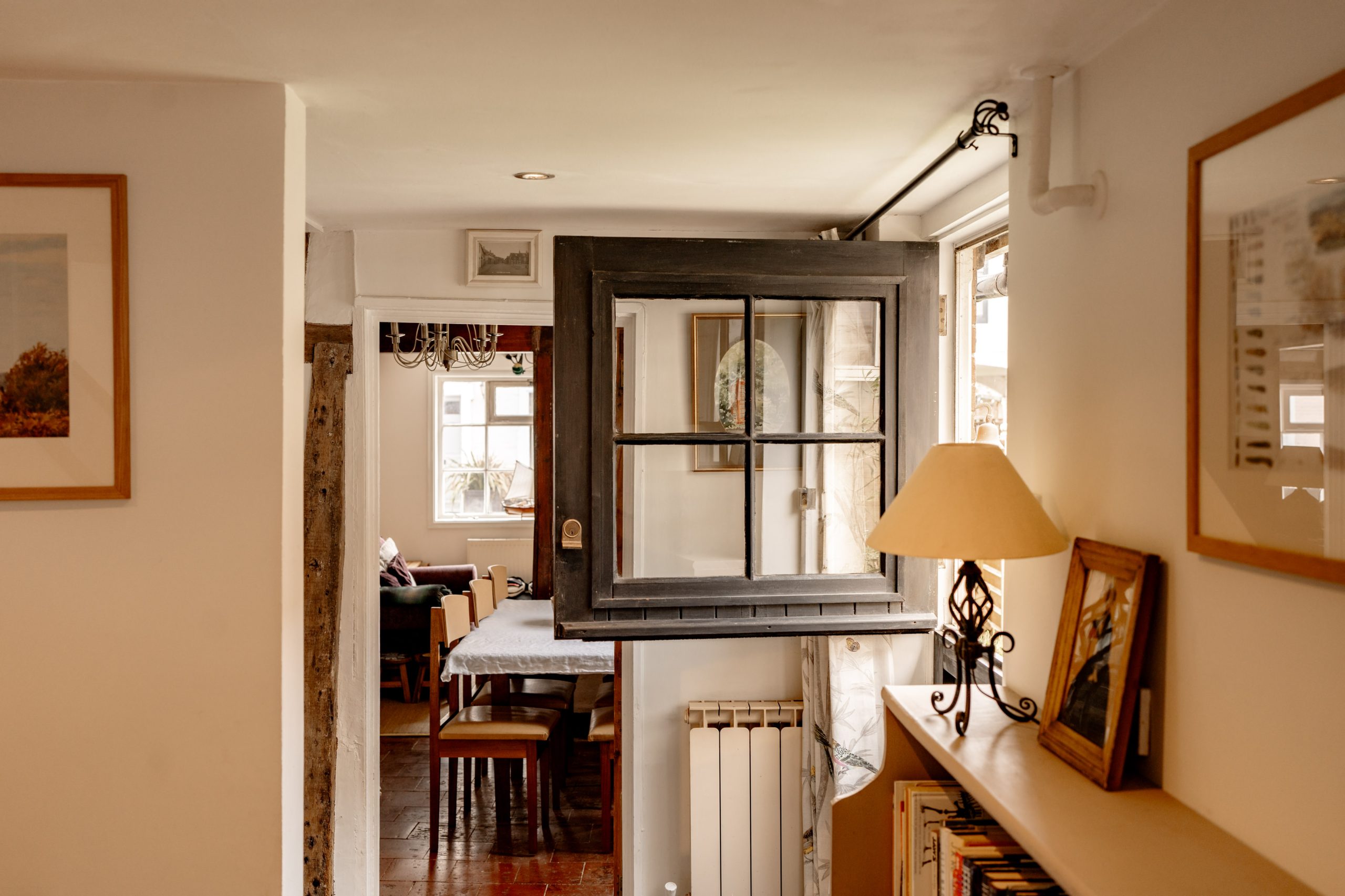 A cozy, rustic interior features a small dining area viewed through a vintage black-framed window mounted inside the house. The room is decorated with framed pictures, a table with chairs, a chandelier, and shelves holding books and a lamp. Natural light fills the space.