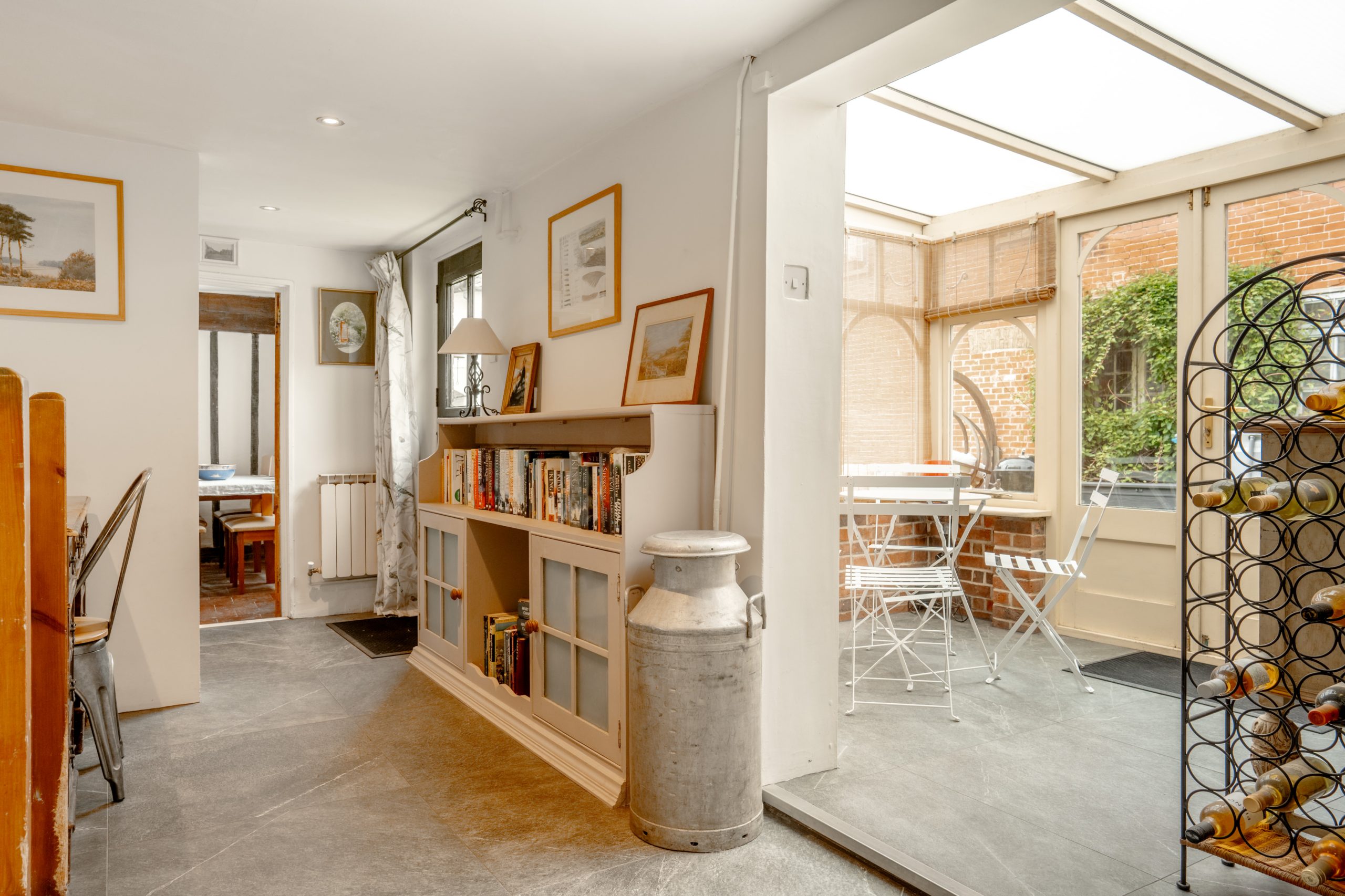 A bright, modern kitchen with grey flooring and white walls. A bookshelf holds various books and decorations. A large metal milk can sits nearby. In the background, there is a sunlit room with a glass ceiling, housing a white table and chairs.