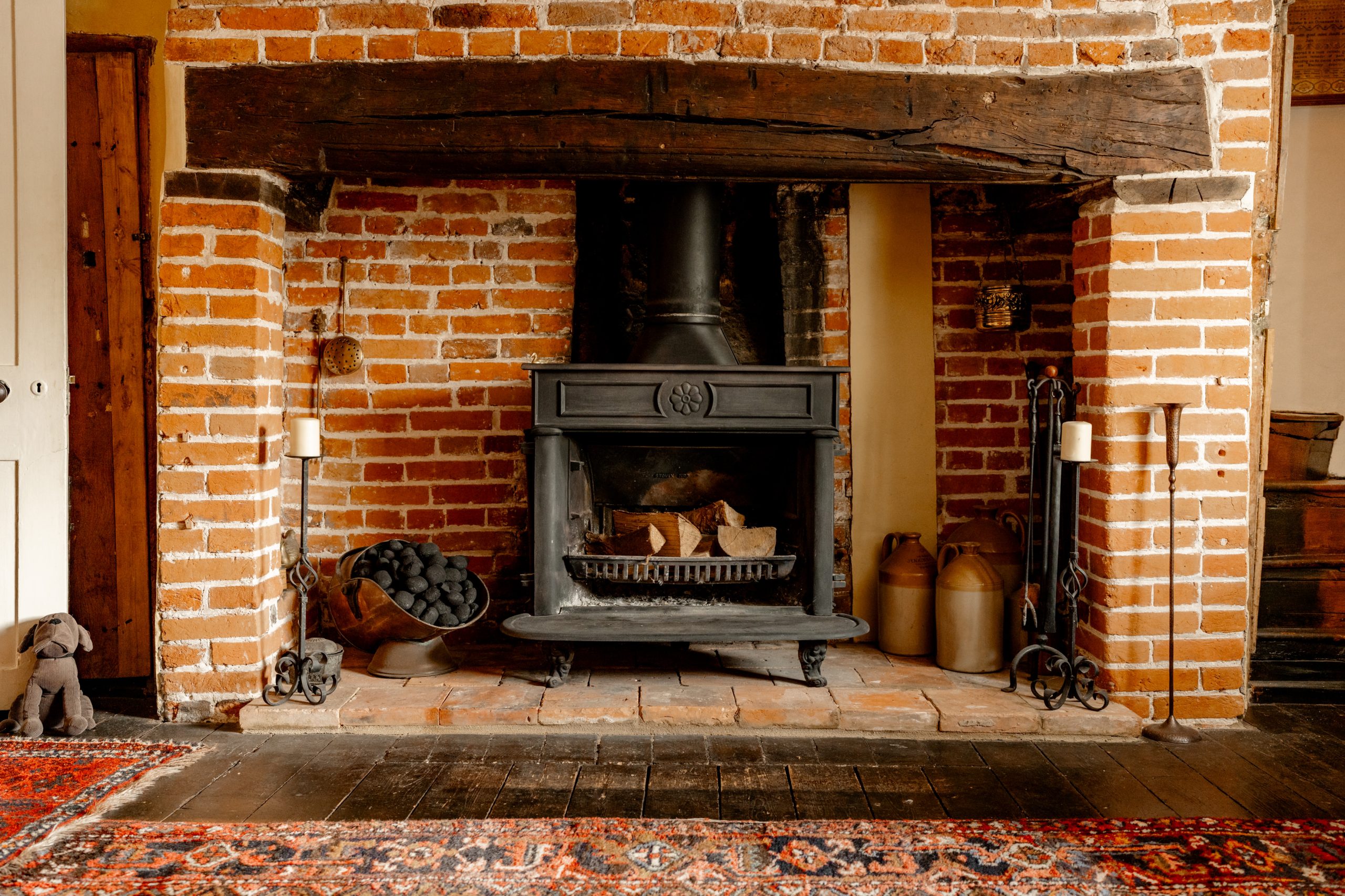A cozy brick fireplace features a black wood-burning stove with firewood inside. A stack of coal sits in a metal basket to the left, flanked by two candles. Two jugs and a metal stand are on the right. A plush dog toy rests on the floor by a patterned rug.