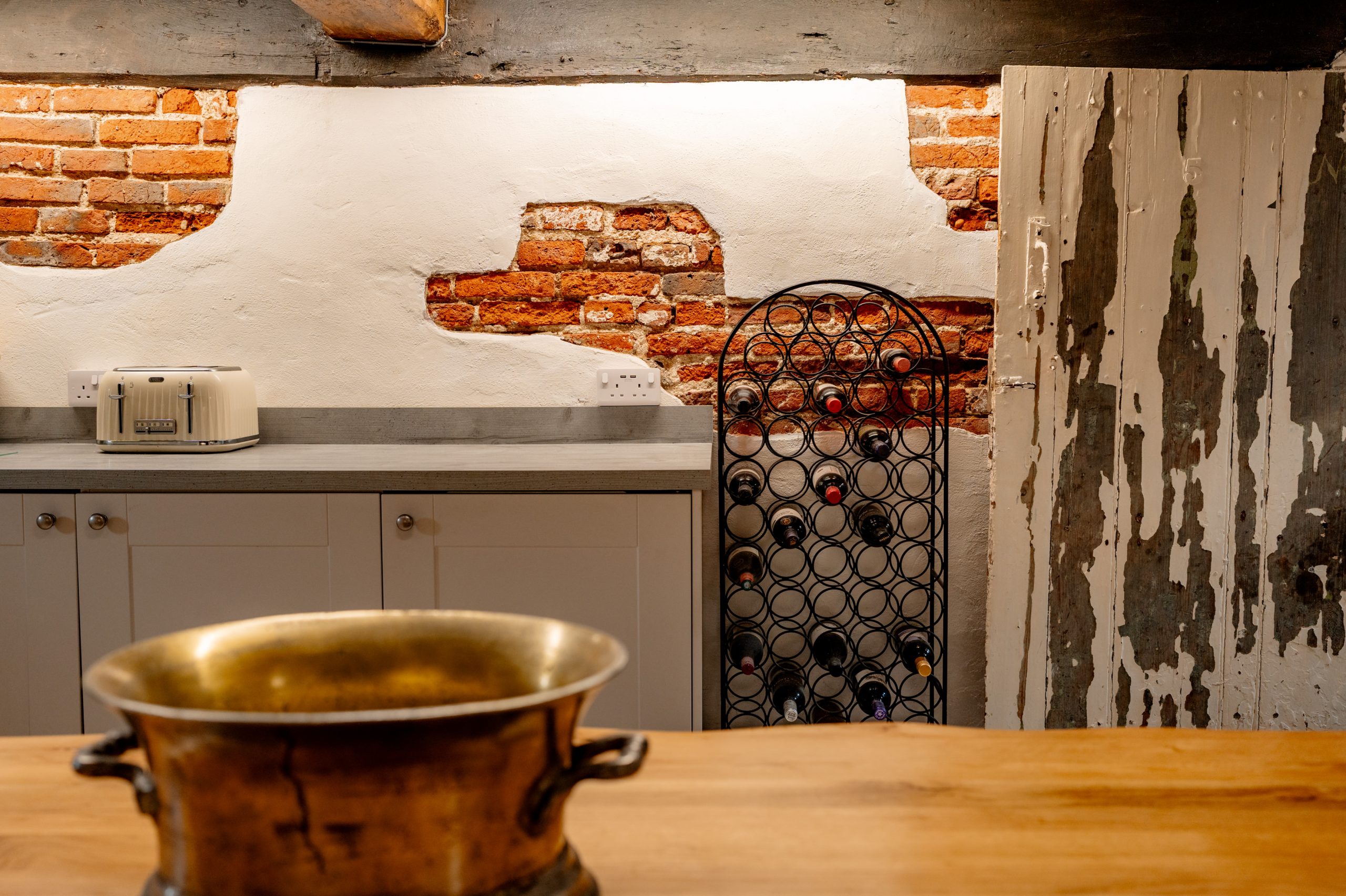 A rustic kitchen features partially exposed brick walls, white cabinets, a light gray countertop, and a wine rack filled with bottles. A toaster sits on the counter, and a weathered wooden door is seen on the right. A brass bowl is on a wooden table in the foreground.