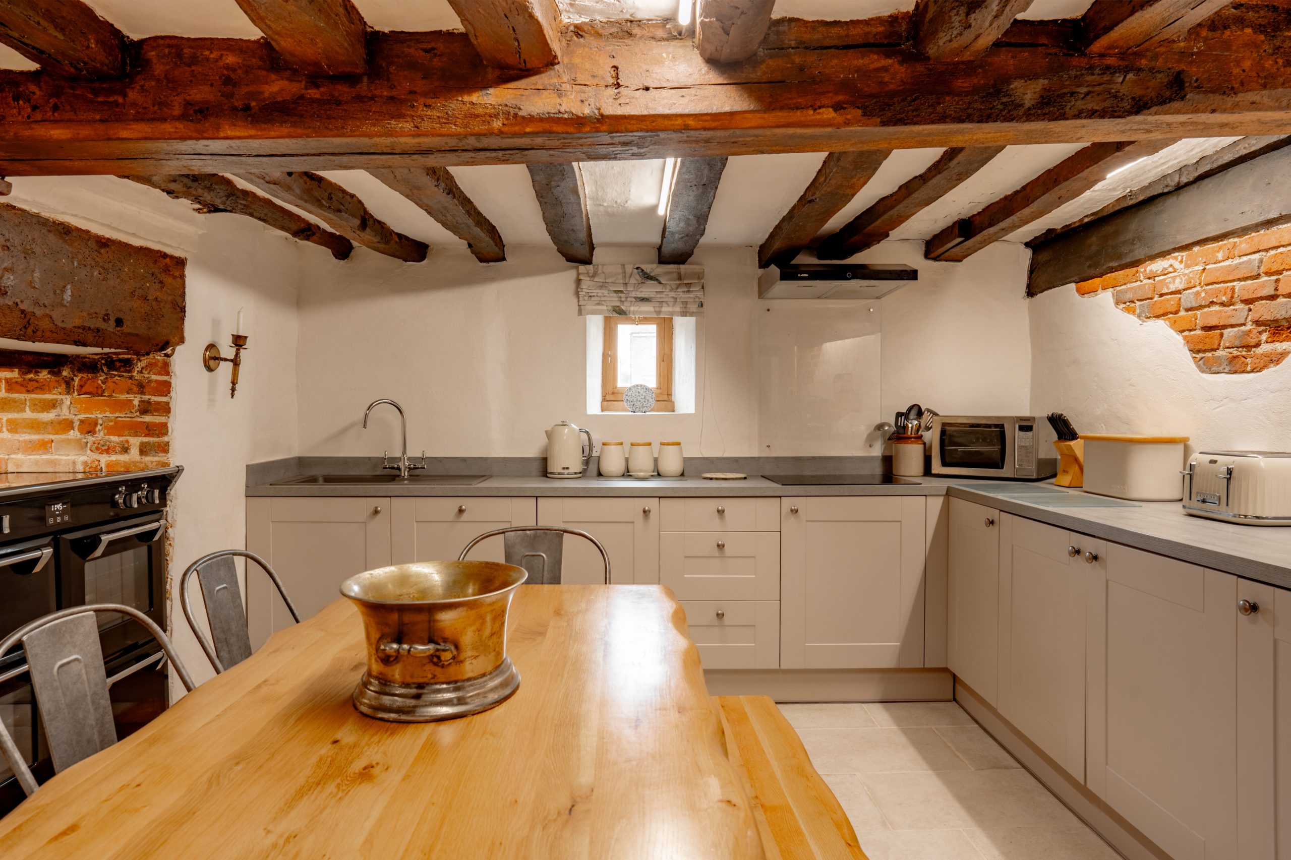 A rustic kitchen with exposed wooden beams and white-washed brick walls. It features modern appliances, including a microwave, toaster, and oven. The room has a large wooden dining table with a metallic bowl centerpiece and metal chairs.