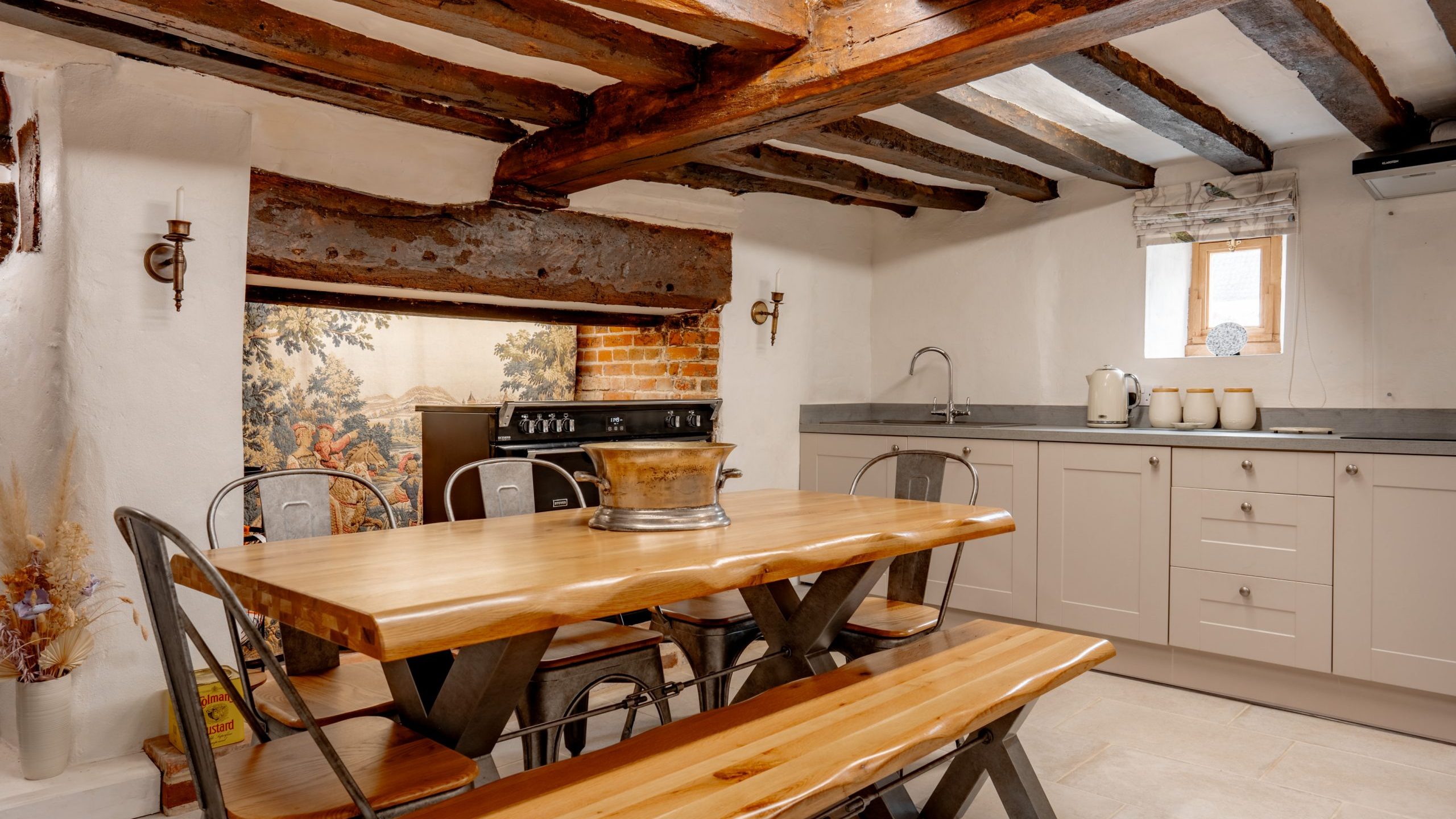 A rustic kitchen with exposed wooden beams features a large wooden dining table with metal chairs and a bench. The kitchen has light-colored cabinets, a countertop with a kettle and jars, a metal sink, and a window allowing natural light to stream in.