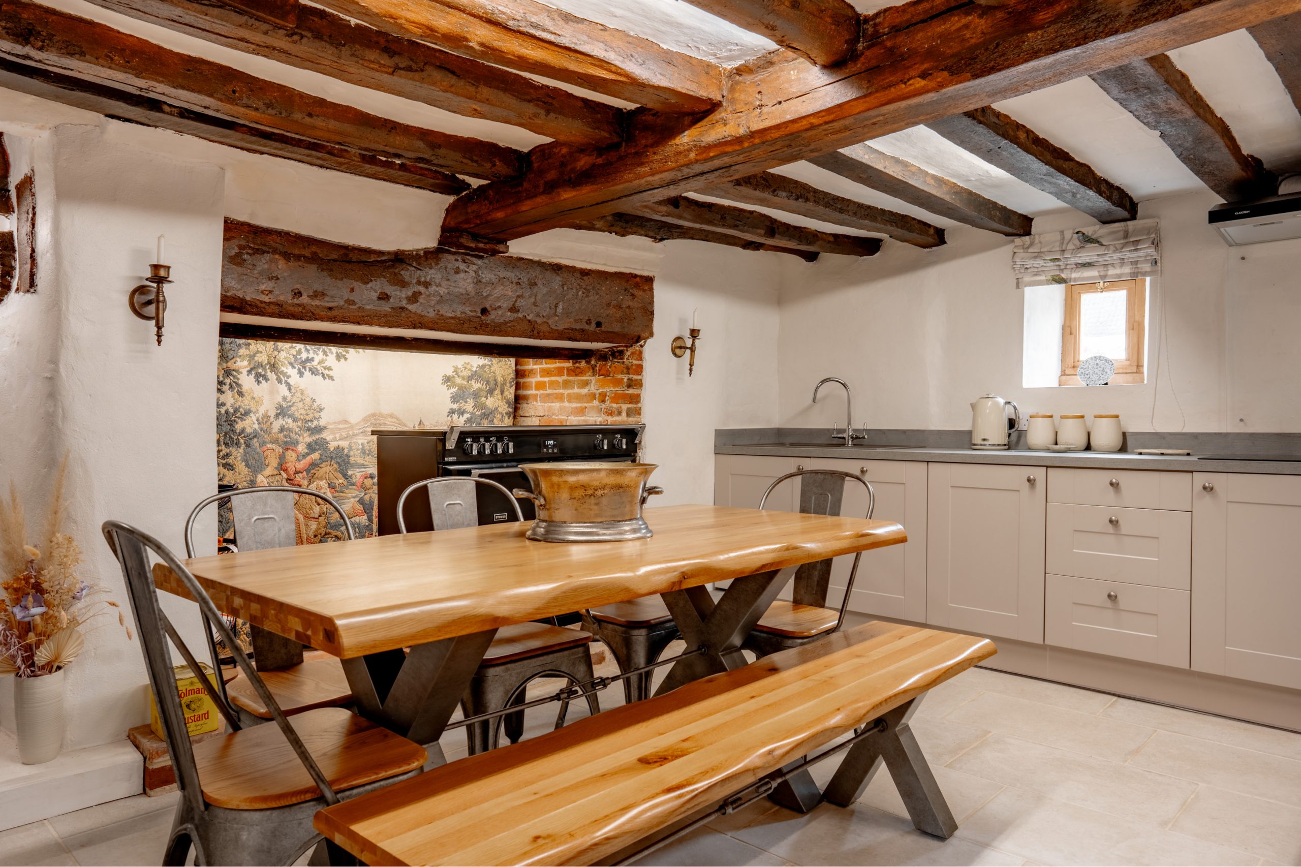 A rustic kitchen with exposed wooden beams features a large wooden dining table with metal chairs and a bench. The kitchen has light-colored cabinets, a countertop with a kettle and jars, a metal sink, and a window allowing natural light to stream in.