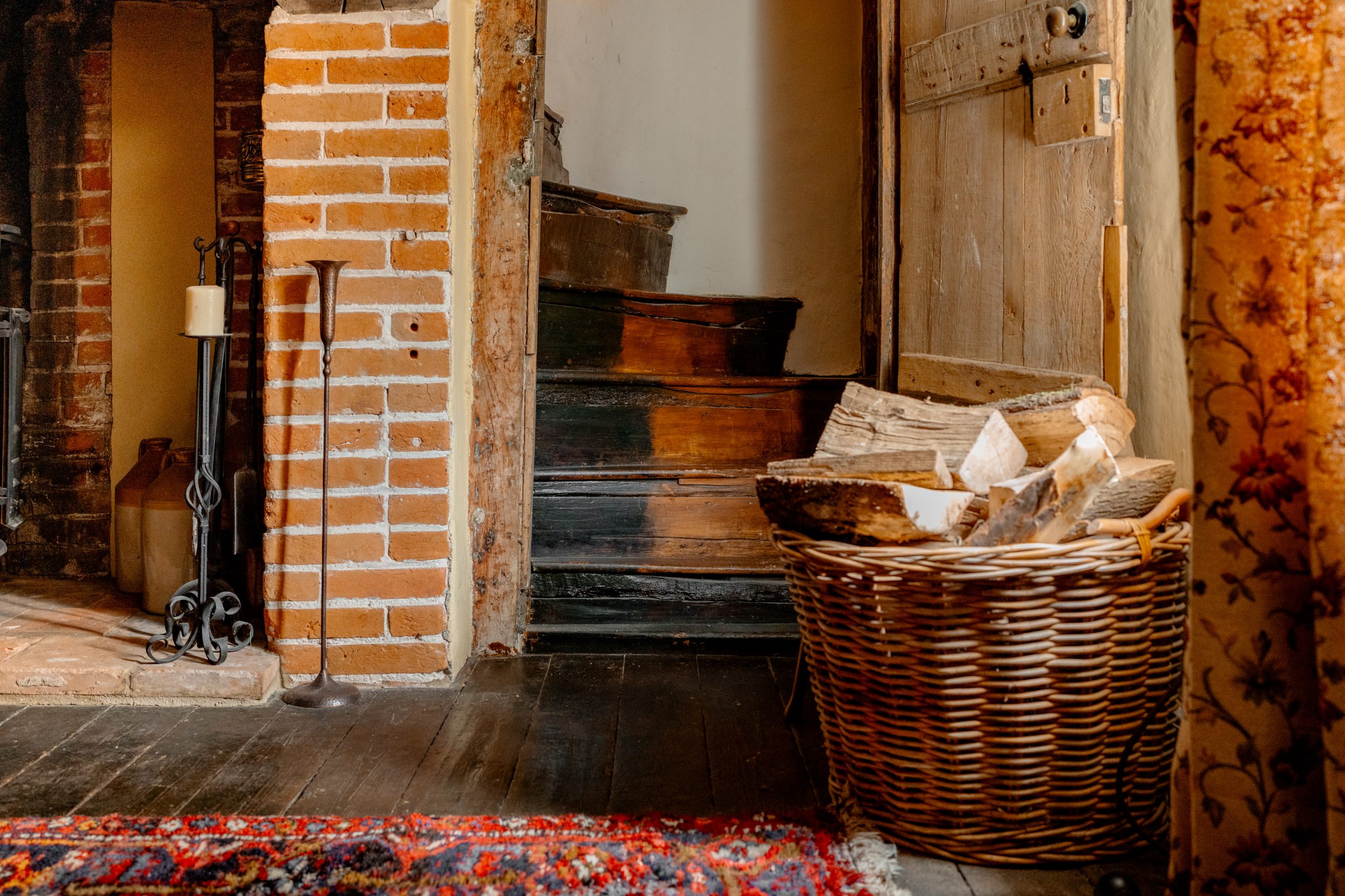 A cozy interior featuring a brick fireplace with black iron tools and a single lit candle on a stand. A wicker basket filled with firewood is placed near an old wooden door. A worn staircase leads up from the rustic wooden floor, partially covered by a patterned rug.