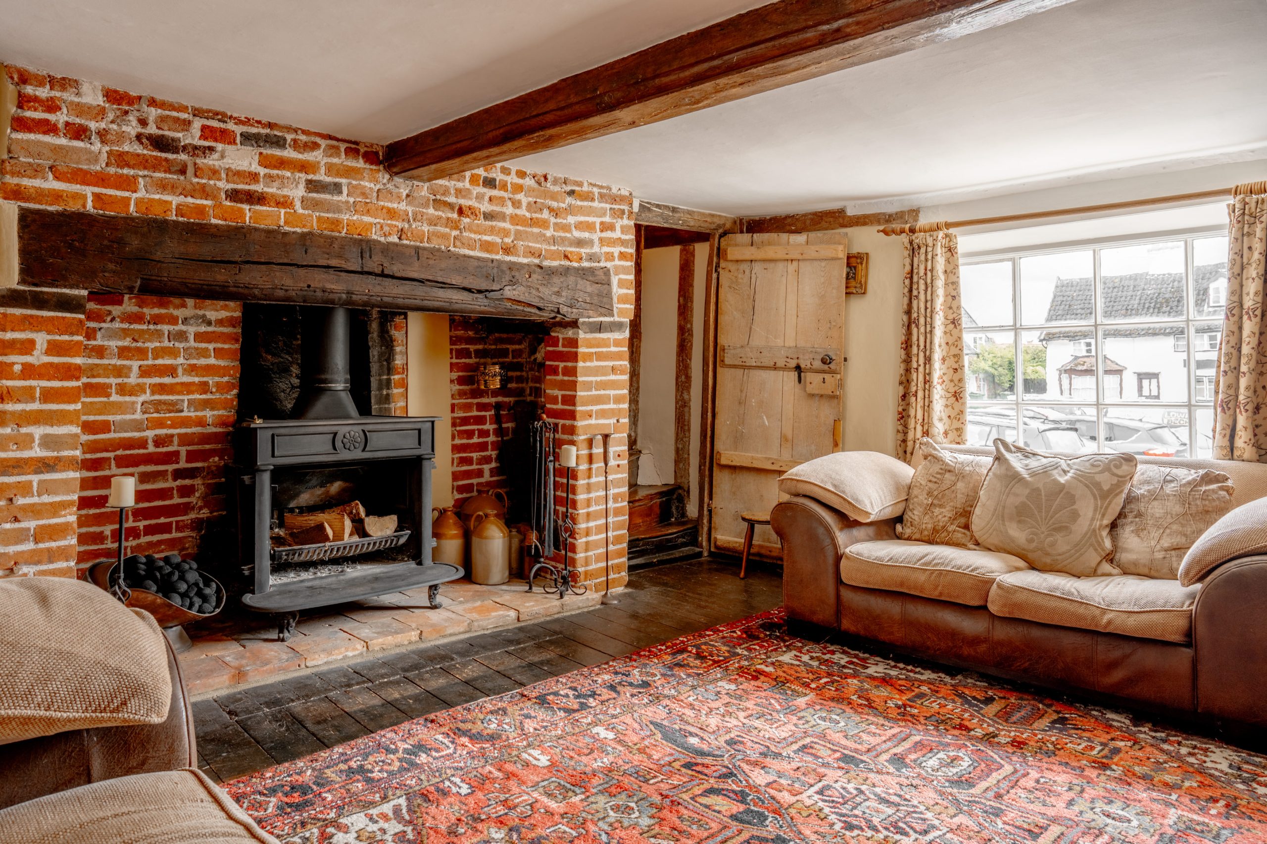 A cozy living room with brick walls and a wood beam ceiling features a fireplace with a black cast iron stove. There are two plush sofas, a patterned red rug, a window with floral curtains, and a wooden door. The room is warmly lit by natural light.