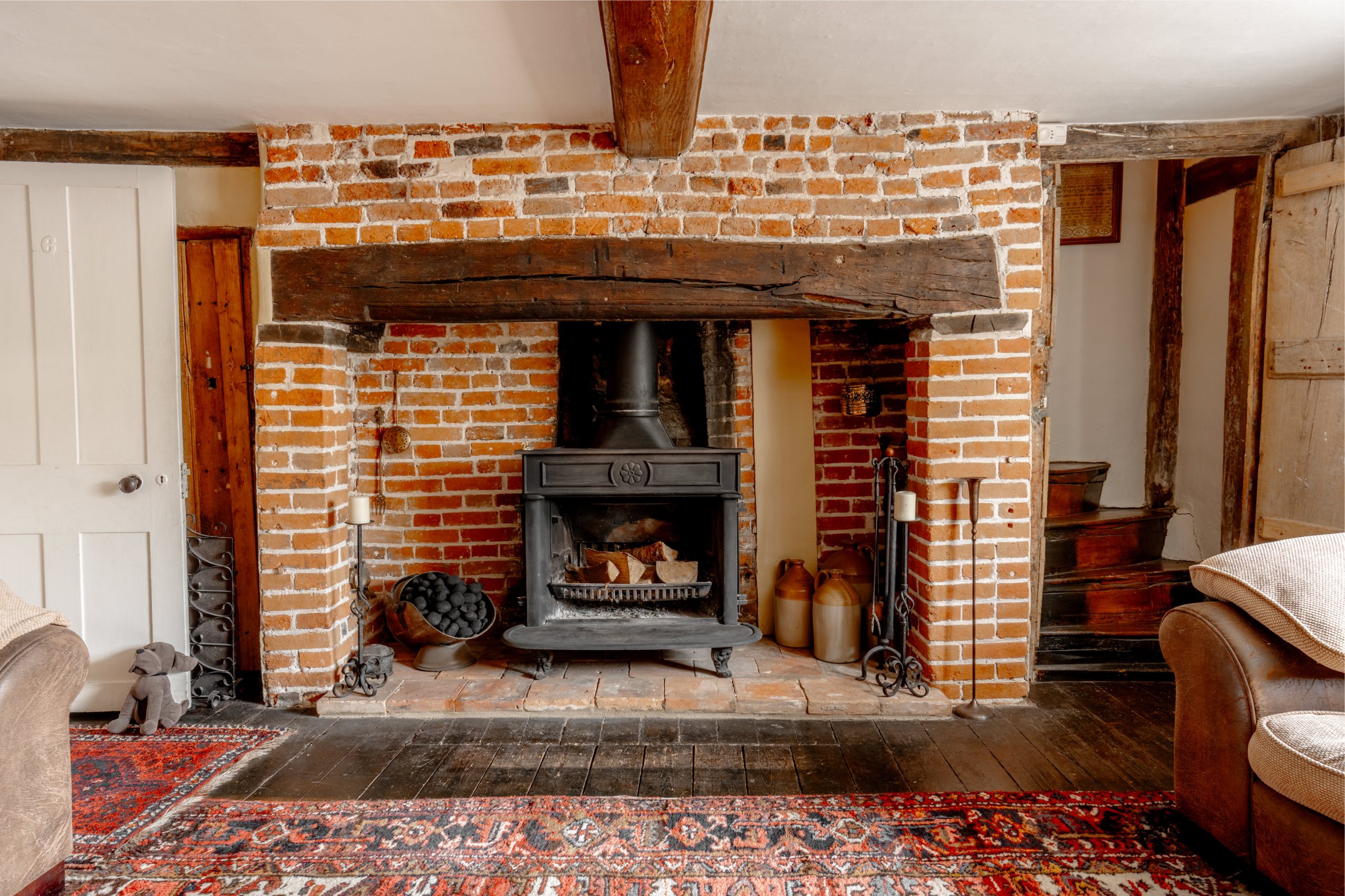 A cozy room featuring a rustic brick fireplace with a black wood-burning stove. Log storage and decorative items flank the fireplace. Exposed wooden beams and a red-patterned area rug add warmth to the space. A doorway and wooden staircase are visible on the sides.
